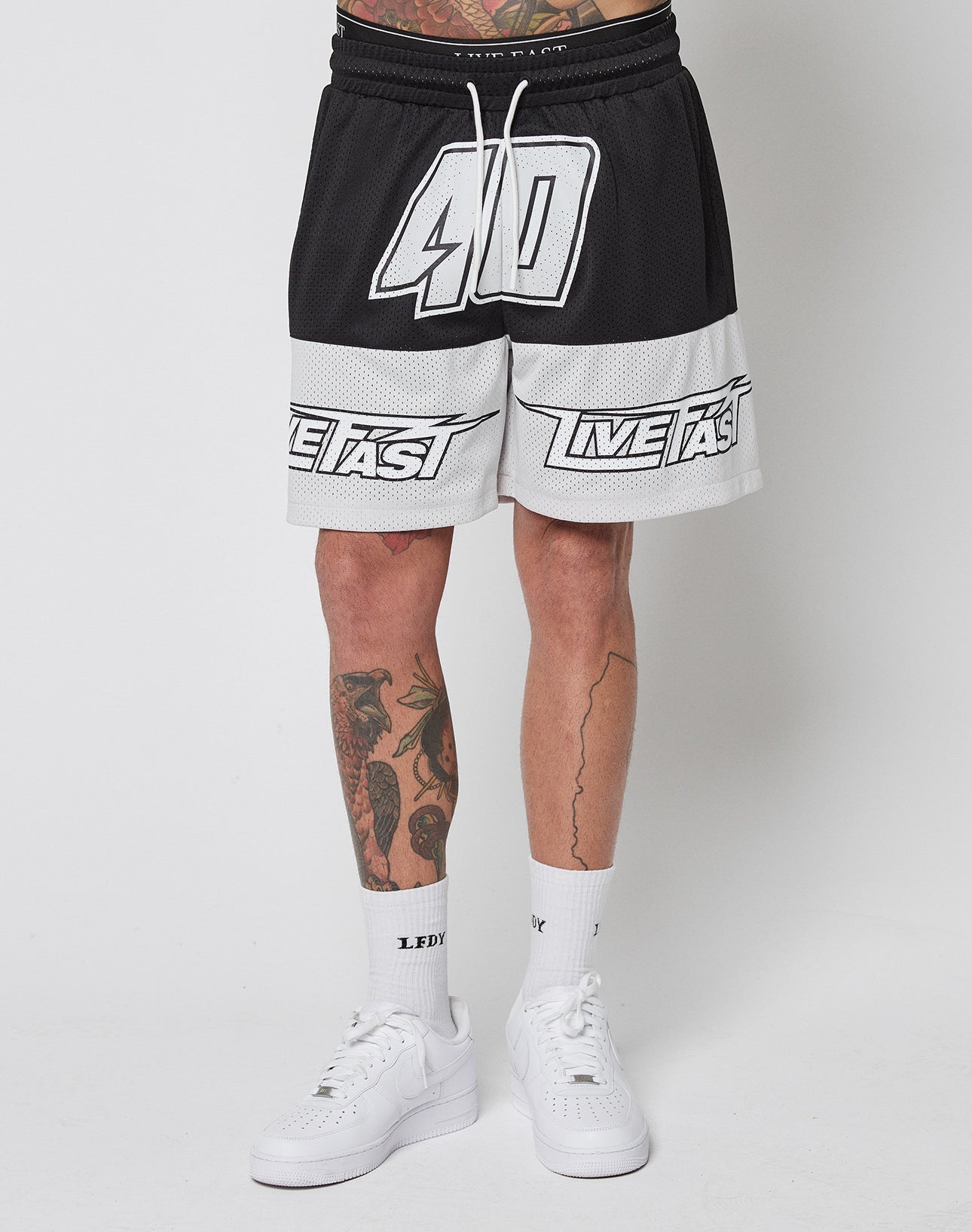 A person wears LFDY Racing Mesh Shorts by LIVE FAST DIE YOUNG—black and white, regular fit, 40 and LIVE FAST printed, elastic waistband, polyester—paired with white crew socks with LEDY text, white sneakers, and visible leg tattoos on a plain background.