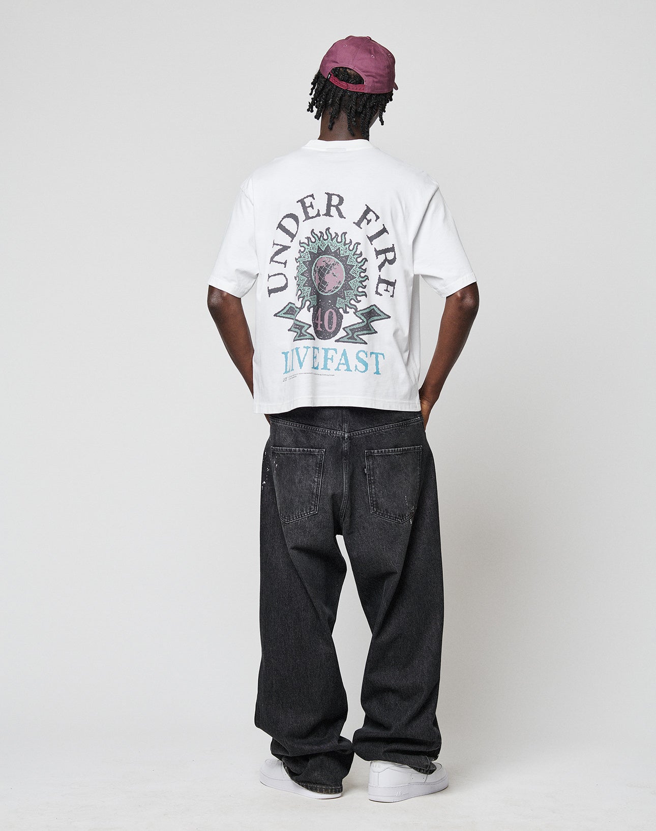Someone wearing the LIVE FAST DIE YOUNG LFDY Under Fire Tee in oversize white, black jeans, white sneakers, and a purple cap stands with their back to the camera against a plain background.