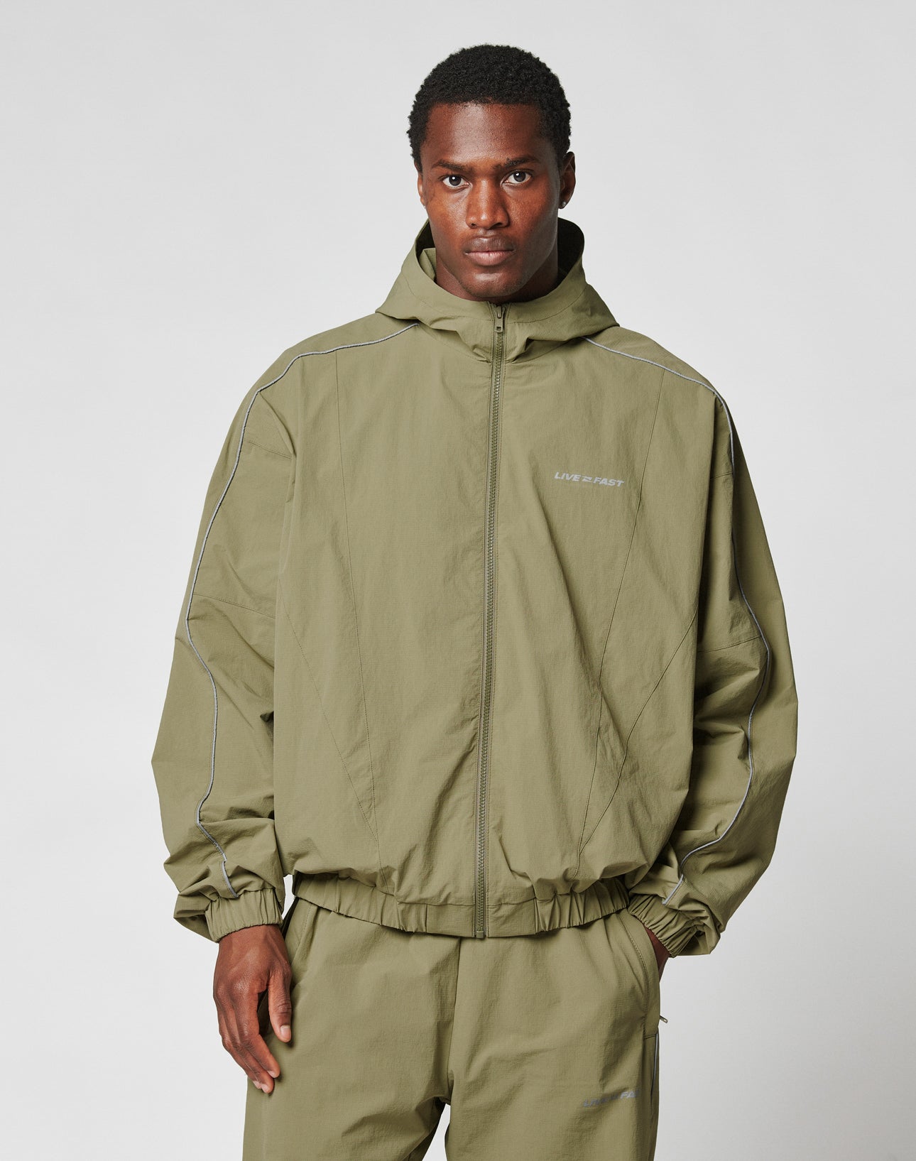 A man faces forward in a plain light gray setting, wearing the olive green Live Fast Trackjacket by LIVE FAST DIE YOUNG. The nylon jacket has a zipper, hood, reflektierende Pipings, subtle white piping, matching pants, and a small chest logo.