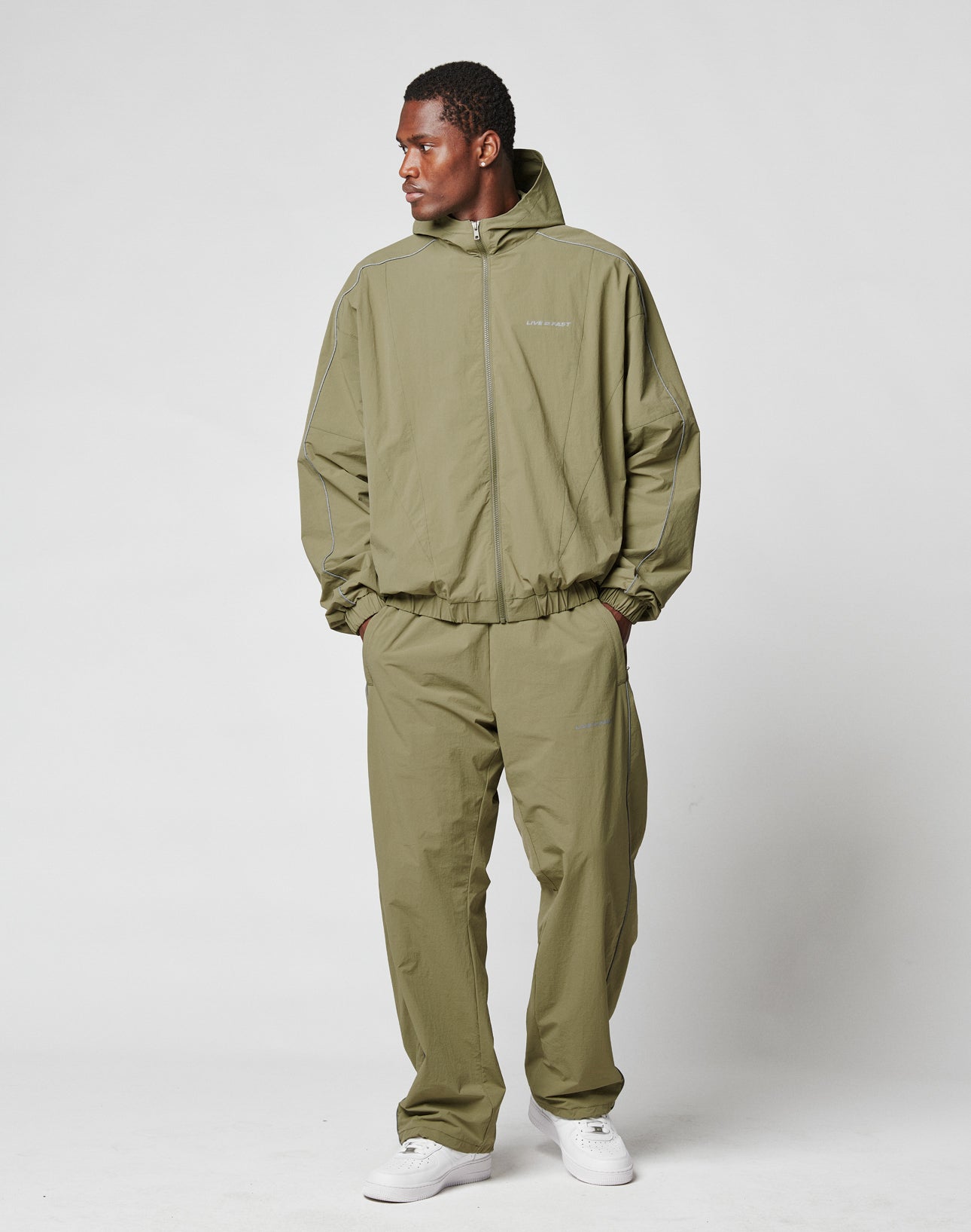 A man stands against a plain background wearing LIVE FAST DIE YOUNG’s olive Live Fast Trackpants and matching hooded top with Elastik-Kordelzug. The wide fit is paired with white sneakers as he looks left, hands in his pockets.