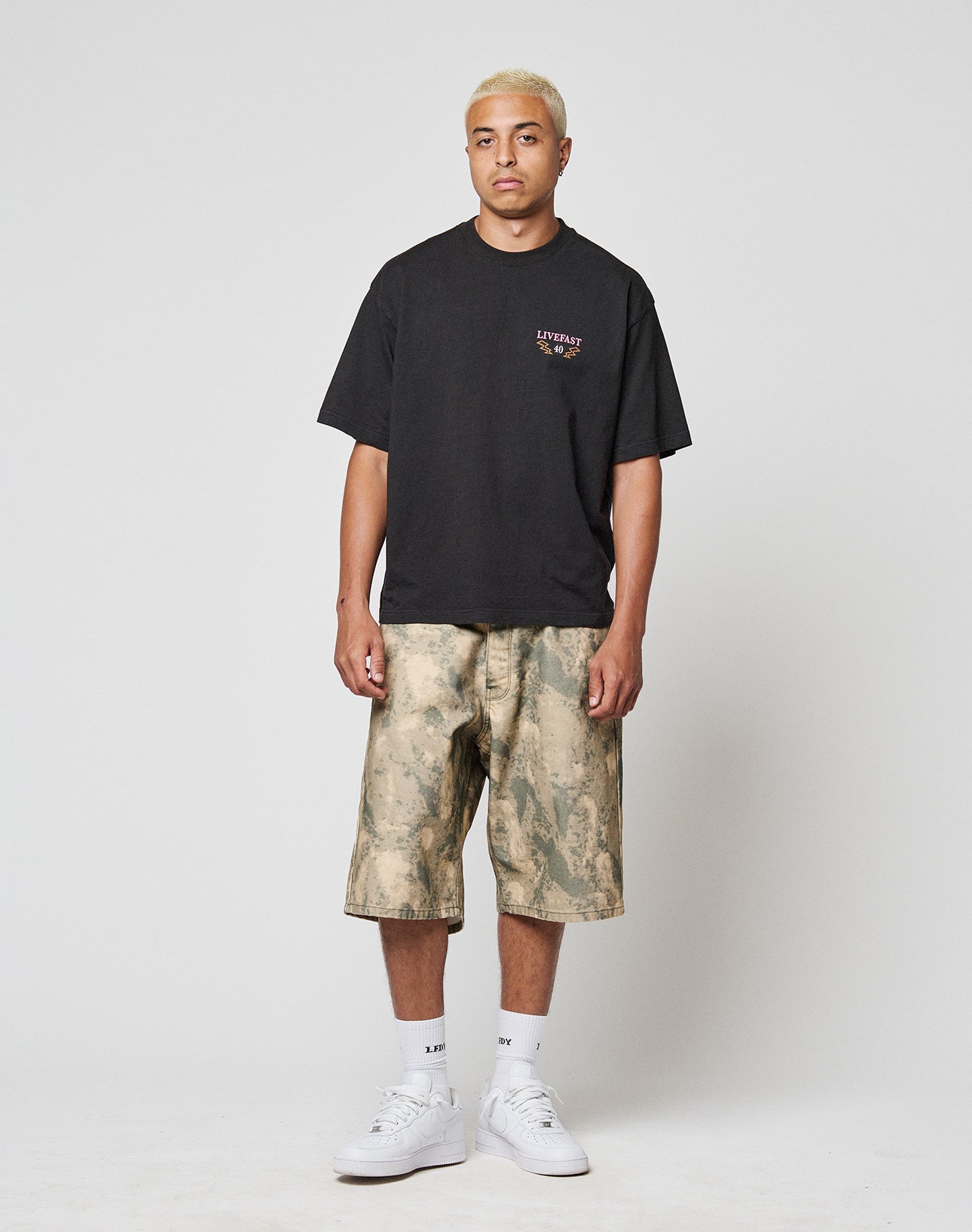 A man with short blond hair wears a baggy black T-shirt, white crew socks, white sneakers, and LIVE FAST DIE YOUNG Canvas Shorts—camouflage-patterned, knee-length, 100% Baumwolle—standing against a plain white background.