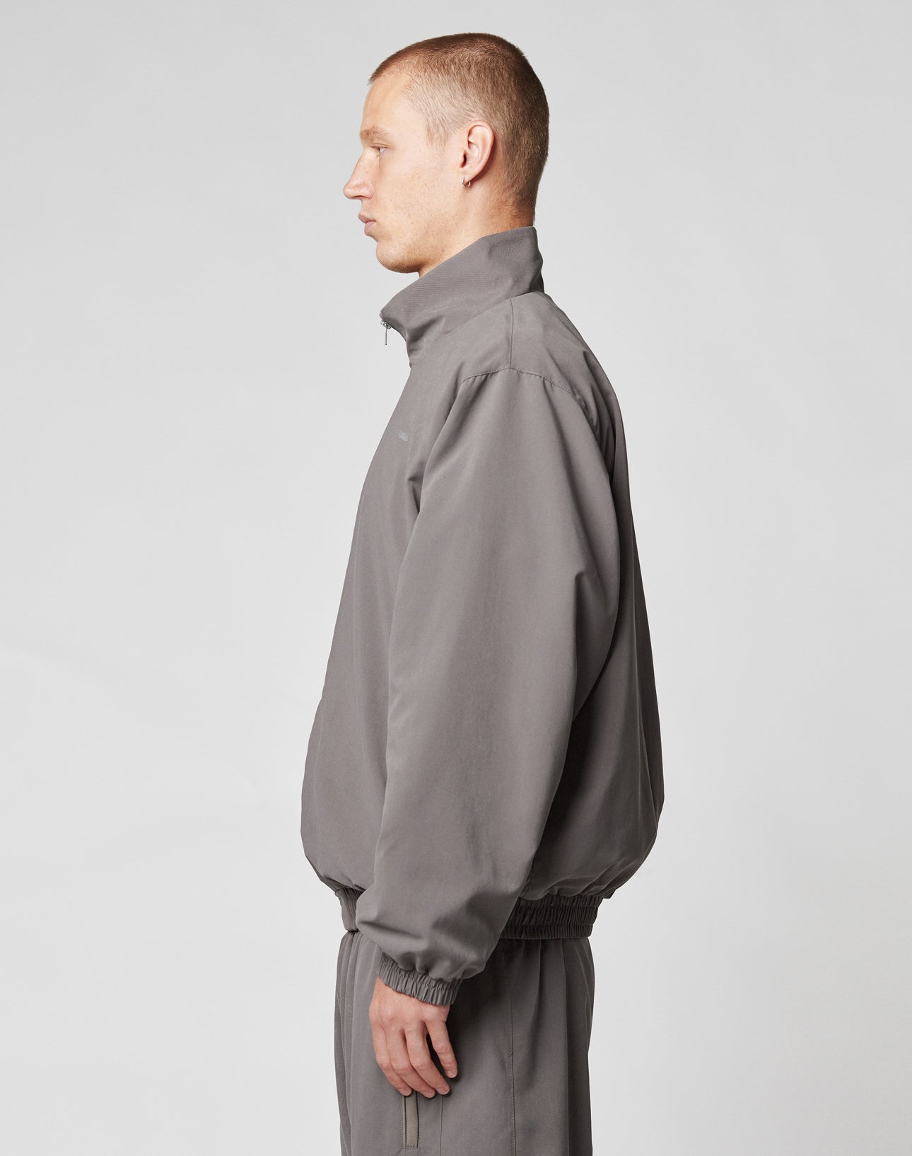 A man with short hair stands in profile, facing left, wearing a light gray LFDY Trackjacket and matching pants from LIVE FAST DIE YOUNG with an oversize fit and reflective print, set against a plain light background.