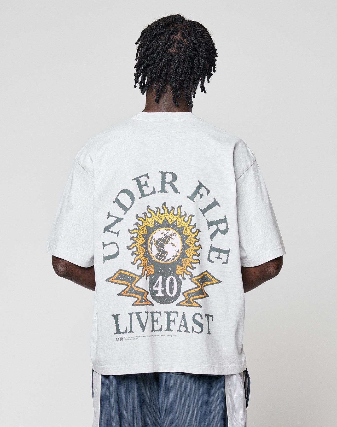 A person faces away, wearing the LIVE FAST DIE YOUNG LFDY Under Fire Tee in light gray, featuring a globe, sun, and lightning bolts with UNDER FIRE, 40, and LIVE FAST—ideal for fans of statement front and back graphics.