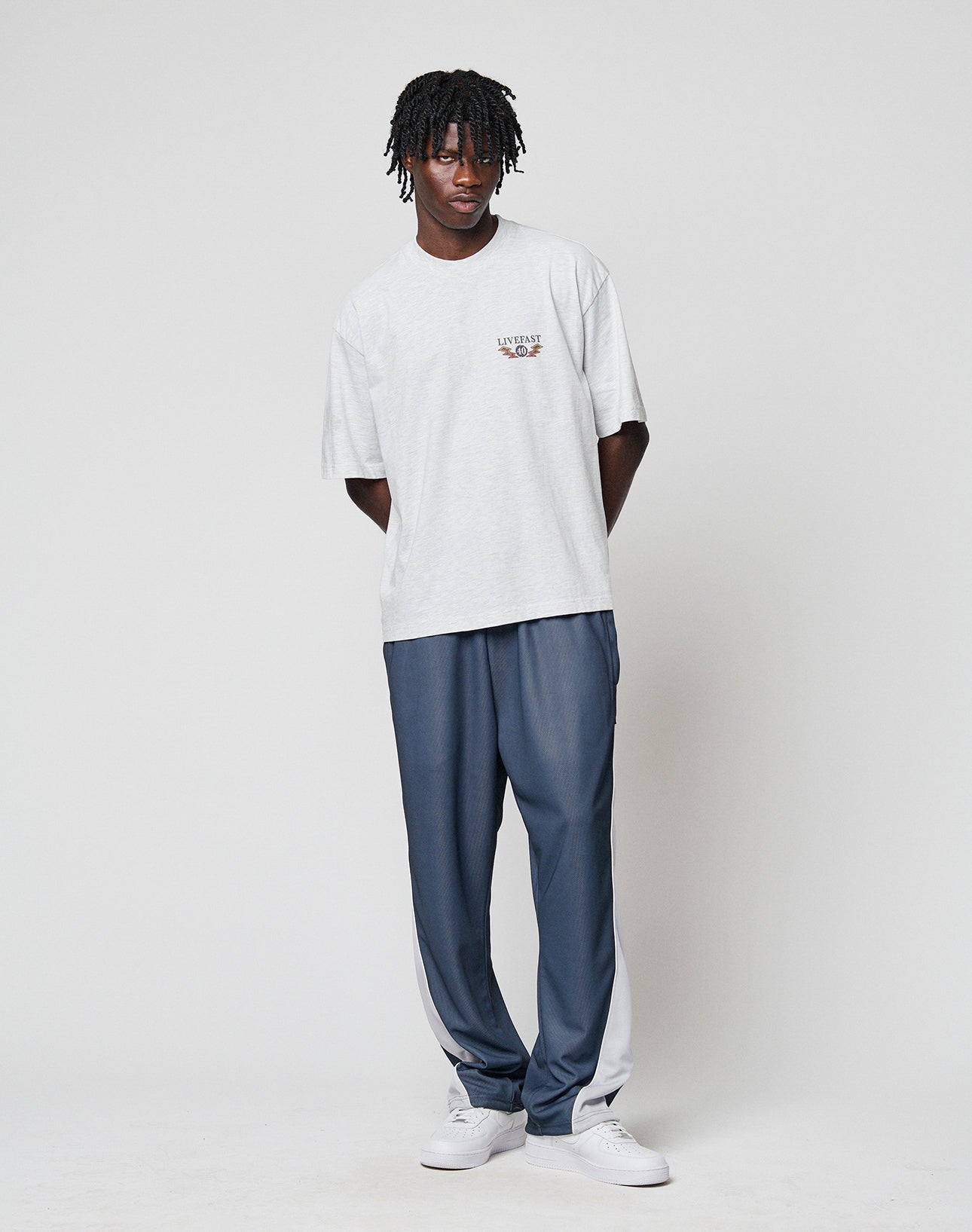 A man with short, twisted hair stands against a white background wearing the LIVE FAST DIE YOUNG LFDY Under Fire Tee in light gray, blue track pants with white side panels, and white sneakers. His hands are behind his back.