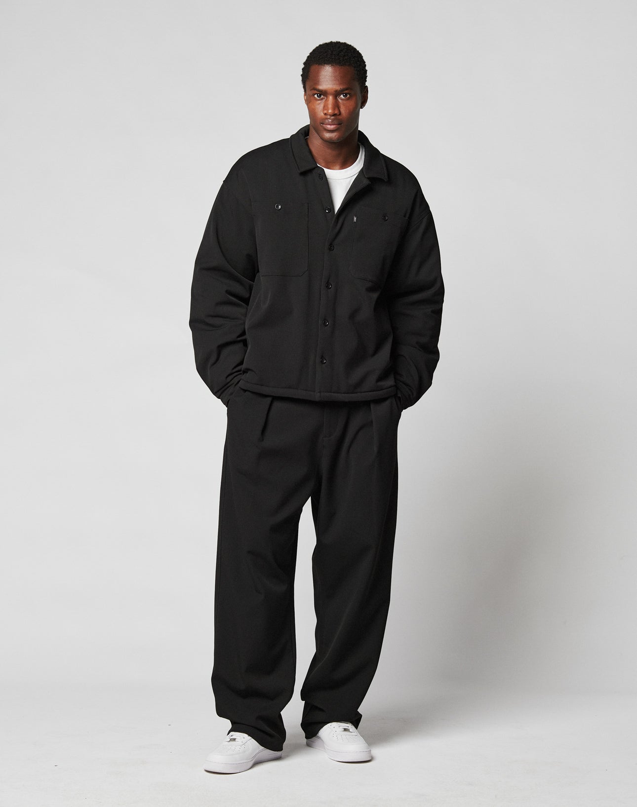 A man stands against a plain light background wearing the LIVE FAST DIE YOUNG Padded Casual Shirt in an oversize fit with chest pockets, matching black pants, and white sneakers over a white tee. His hands are in his pockets as he looks at the camera.