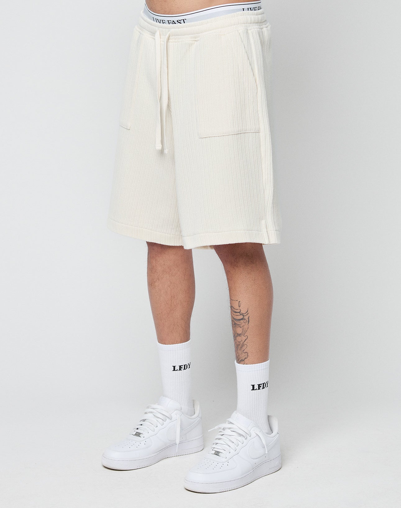 A person wears LIVE FAST DIE YOUNG Basic Knit Shorts with an elastic waistband, white LFDY socks, and white Nike sneakers, standing against a plain light background. Only the lower body and legs are visible.