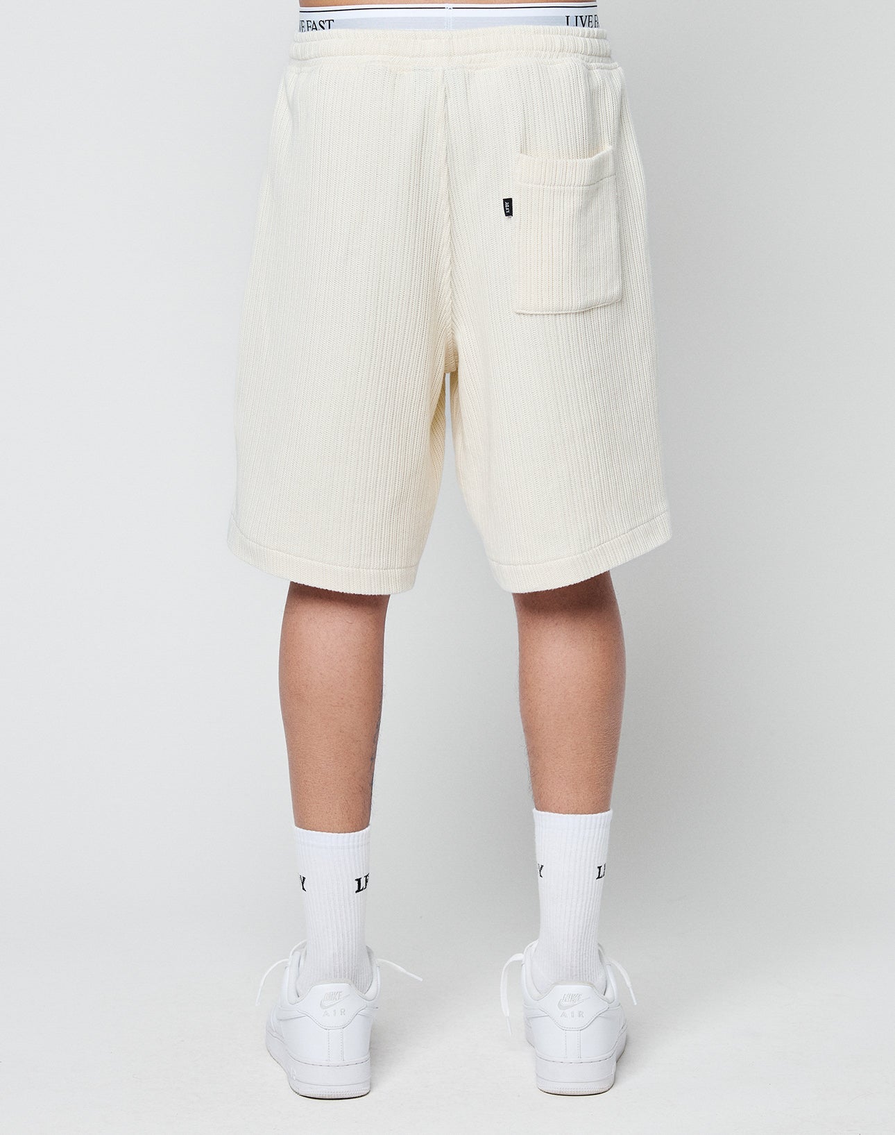 A person is shown from the back wearing off-white LIVE FAST DIE YOUNG Basic Knit Shorts with a back pocket, white crew socks, and white sneakers against a plain light background.
