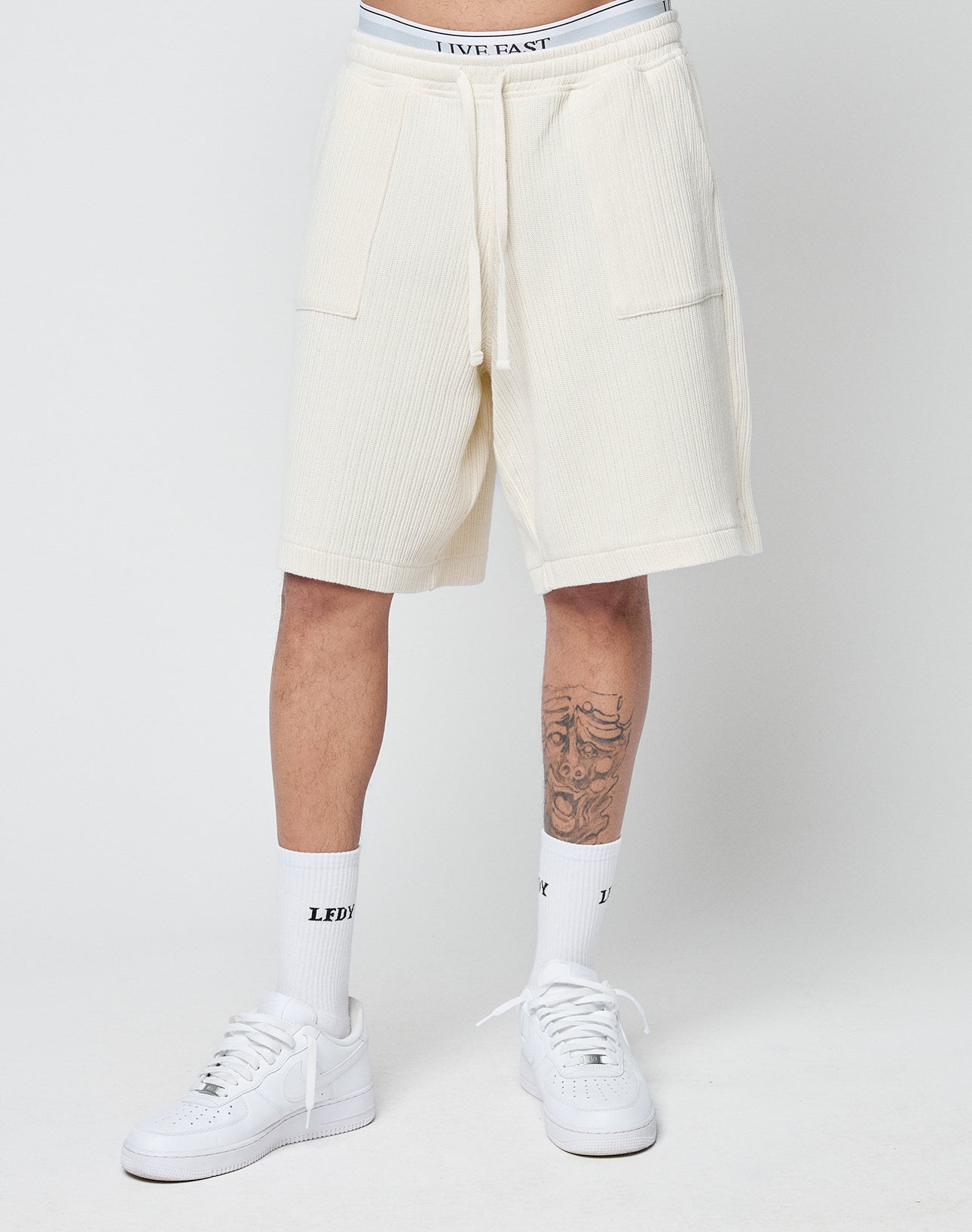 A person wears LIVE FAST DIE YOUNG Basic Knit Shorts in cream with an elastic waistband and patch pockets, white socks with black text, and white sneakers. The lower half is shown against a plain background, revealing a leg tattoo.