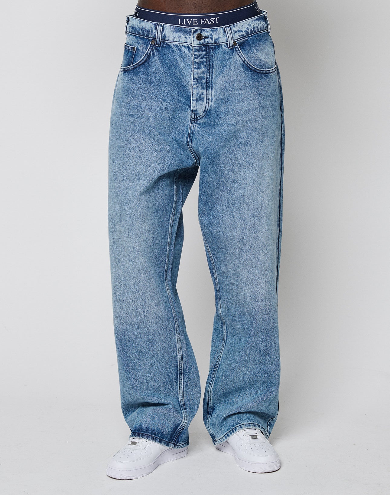 A person wears LFDY Baggy Denim by LIVE FAST DIE YOUNG—loose-fitting blue jeans with a wide straight leg—paired with white sneakers and visible black LIVE FAST DIE YOUNG underwear, standing against a plain white background.