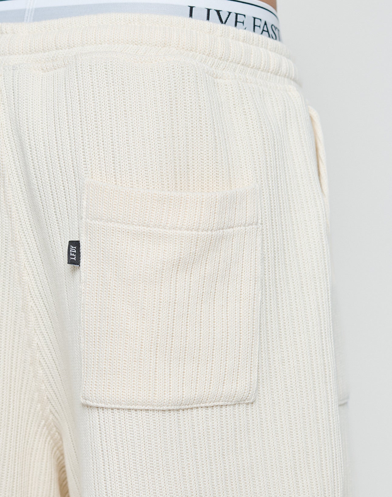 Close-up of cream-colored Basic Knit Shorts by LIVE FAST DIE YOUNG, featuring ribbed knit, patch pockets, and a small black tag. The elastic waistband of underwear with the text LIVE FAST is visible above the shorts.