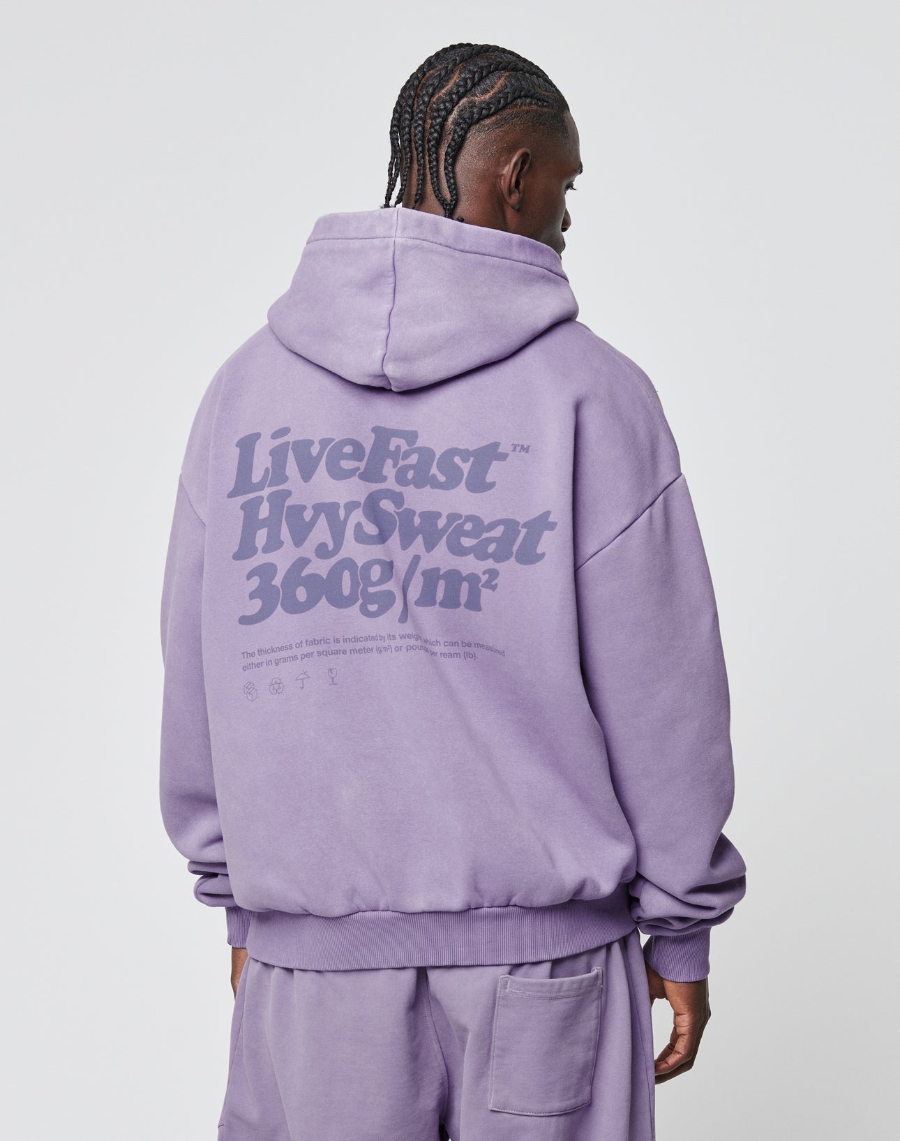A person with braided hair wears an oversize fit, loose lavender Basic 360 Hooded and matching pants by LIVE FAST DIE YOUNG. Bold LiveFast HvySweat 360g/m² text, plus smaller details, appear on the back. Made from premium cotton-polyester.