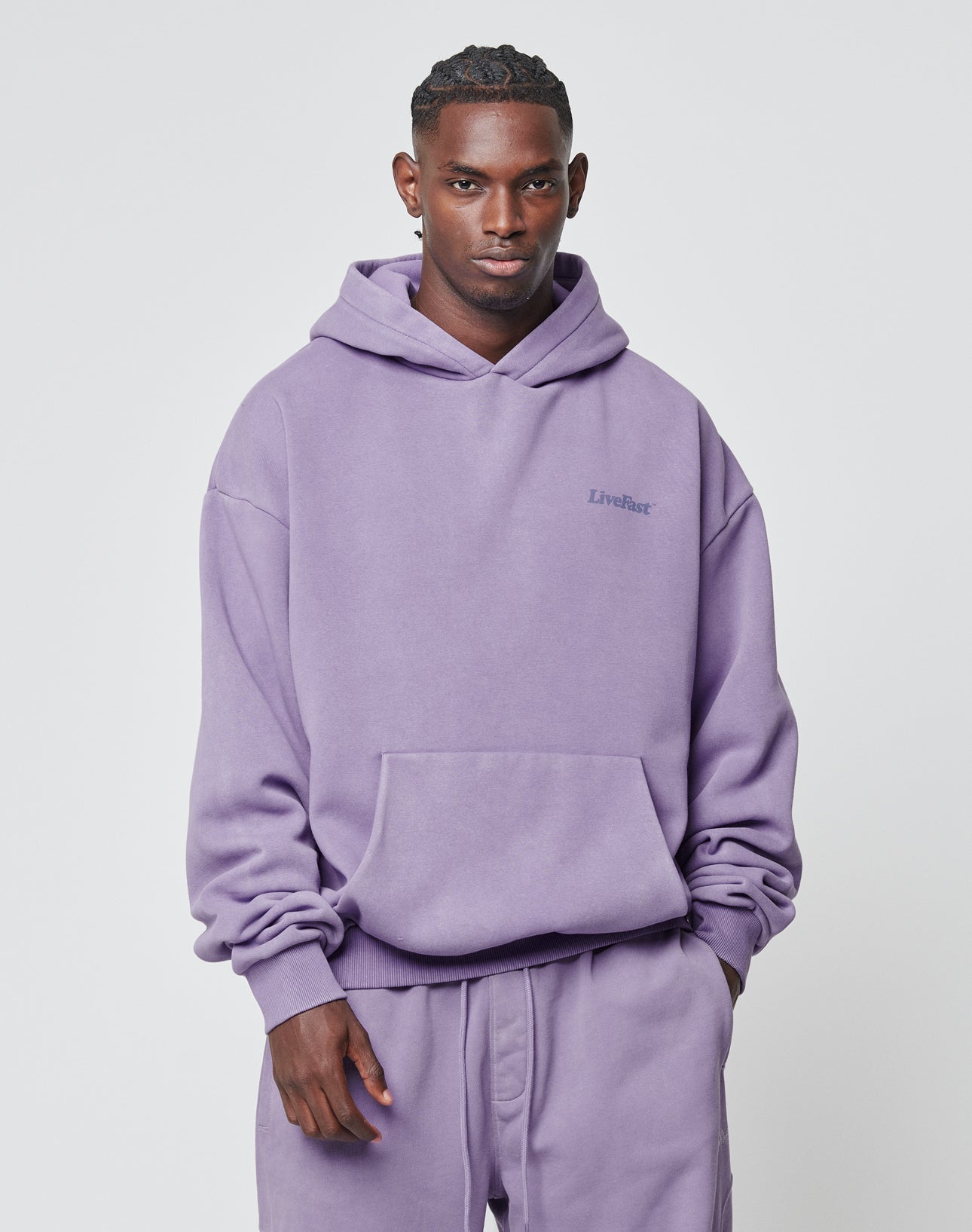 A man faces forward in a loose, lavender LIVE FAST DIE YOUNG Basic 360 Hooded and matching sweatpants, both in Oversize Fit. The hoodie includes a front pocket, left chest text, and soft Baumwolle-Polyester blend. Plain, light background.