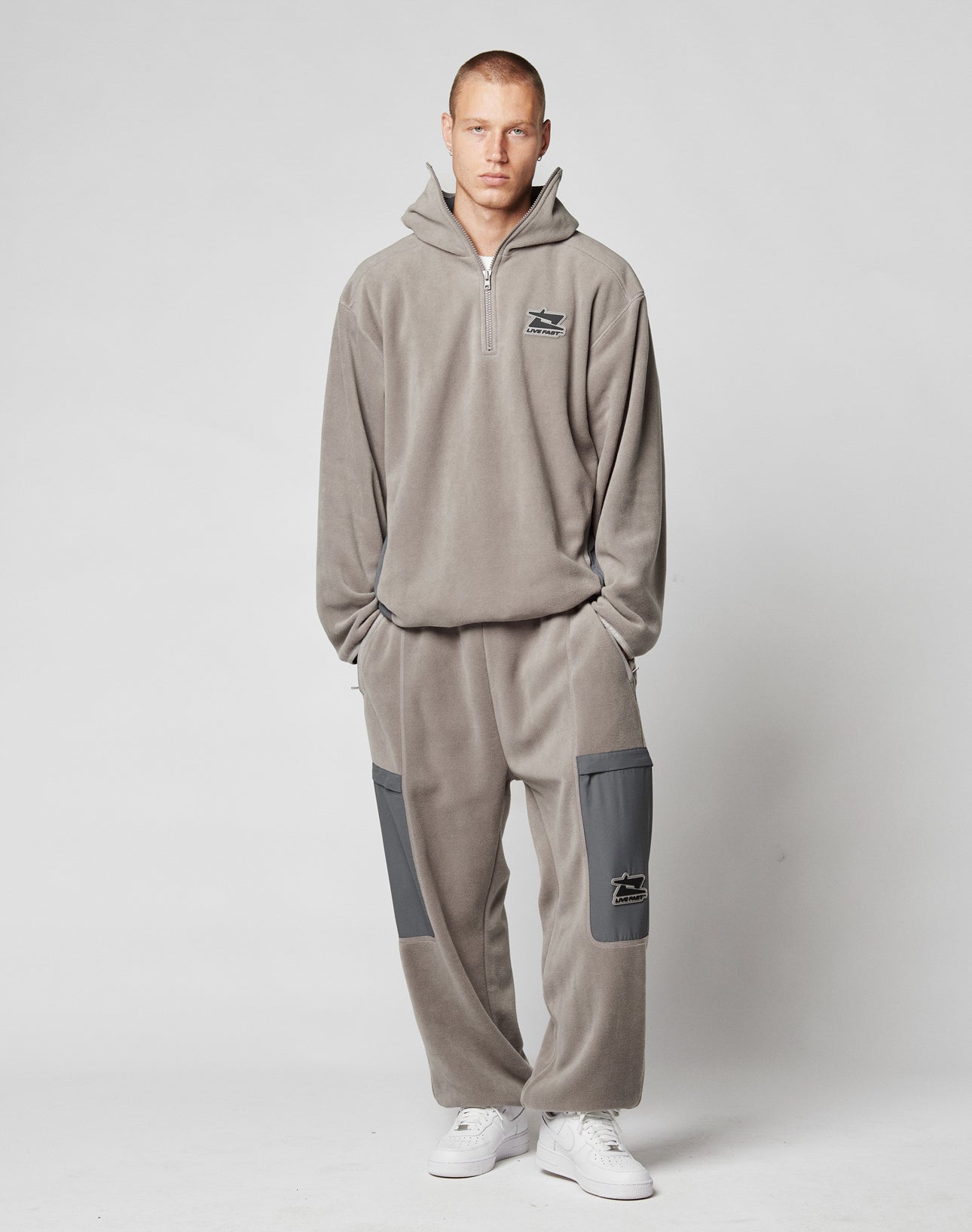 A man with short hair stands facing forward, wearing LIVE FAST DIE YOUNG’s Live Fast Fleece Pants—a loose-fitting light gray tracksuit pant with a wide fit, nylon pockets, elastic waistband, and white sneakers against a plain backdrop.