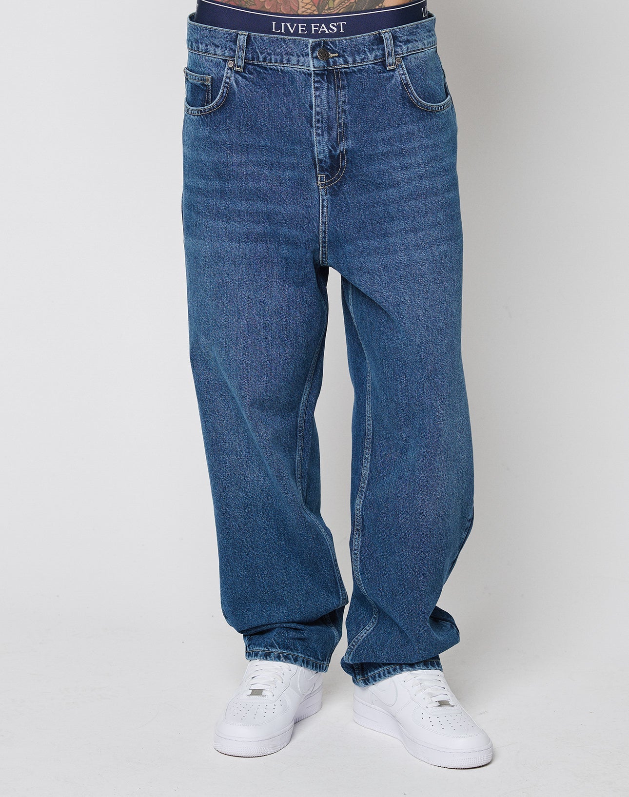 A person stands against a plain light background wearing Wide Denim jeans by LIVE FAST DIE YOUNG, white sneakers, and dark underwear with a visible LIVE FAST DIE YOUNG waistband.