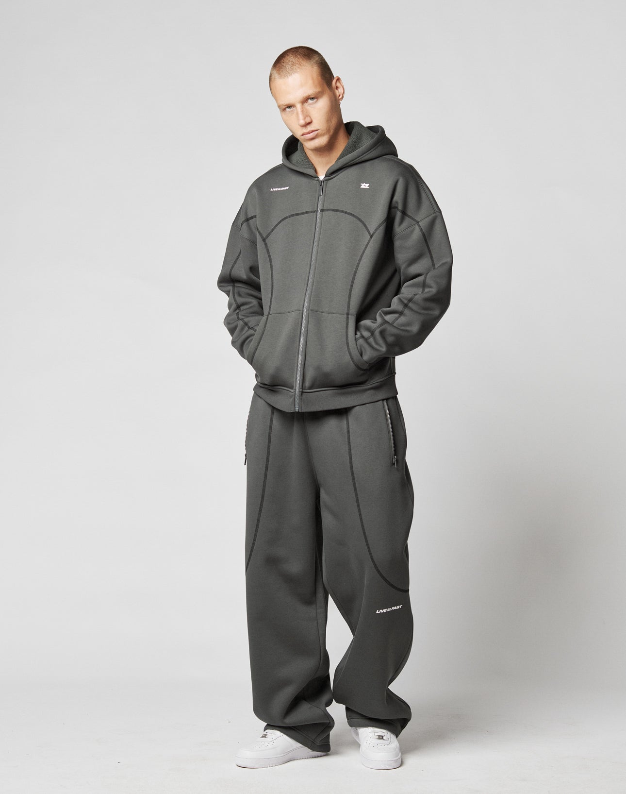 A person wears the LFDY Active Fleece Hooded Zip by LIVE FAST DIE YOUNG with matching baggy sweatpants and white sneakers, standing against a plain light gray background, hands in pockets, looking directly at the camera.