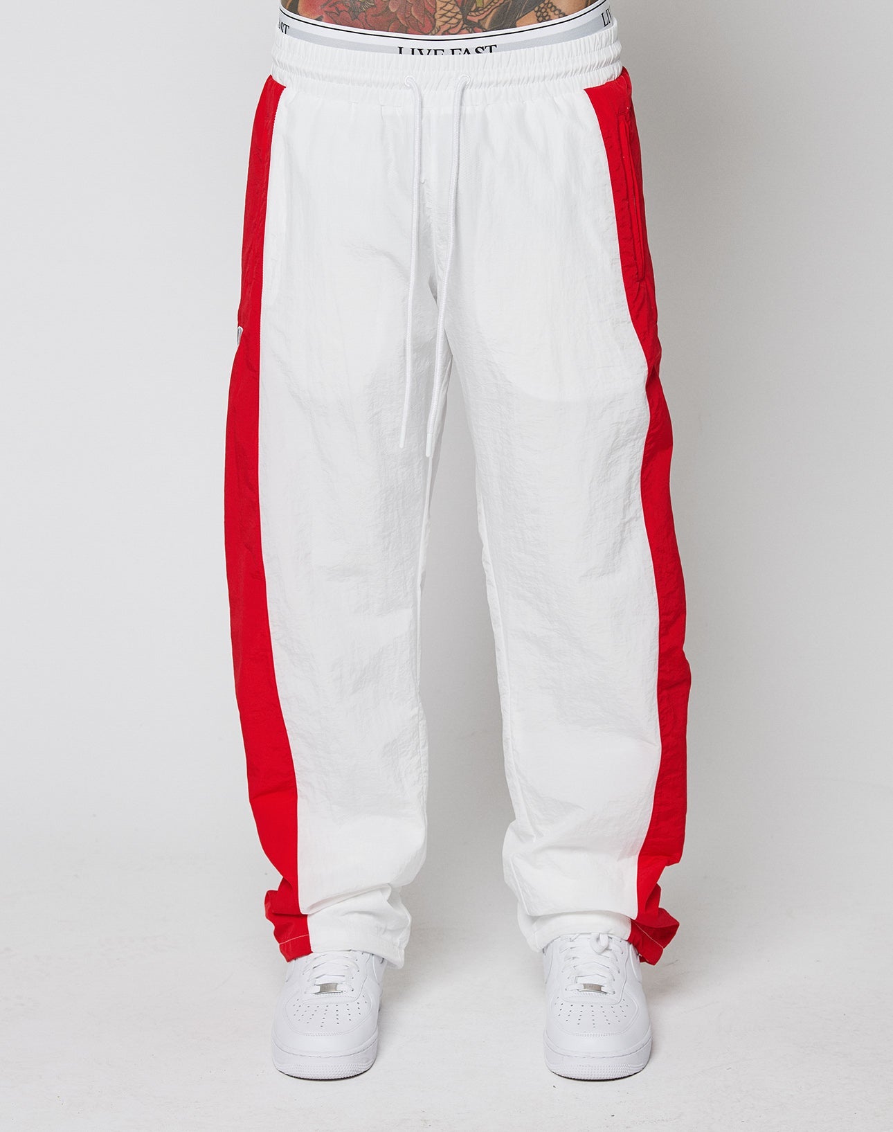 A person wearing LIVE FAST DIE YOUNG LFDY Trackpants 3000—wide fit, white with bold red side stripes, elastic waistband, and crafted from lightweight polyamide—pairs them with white sneakers against a plain white background.