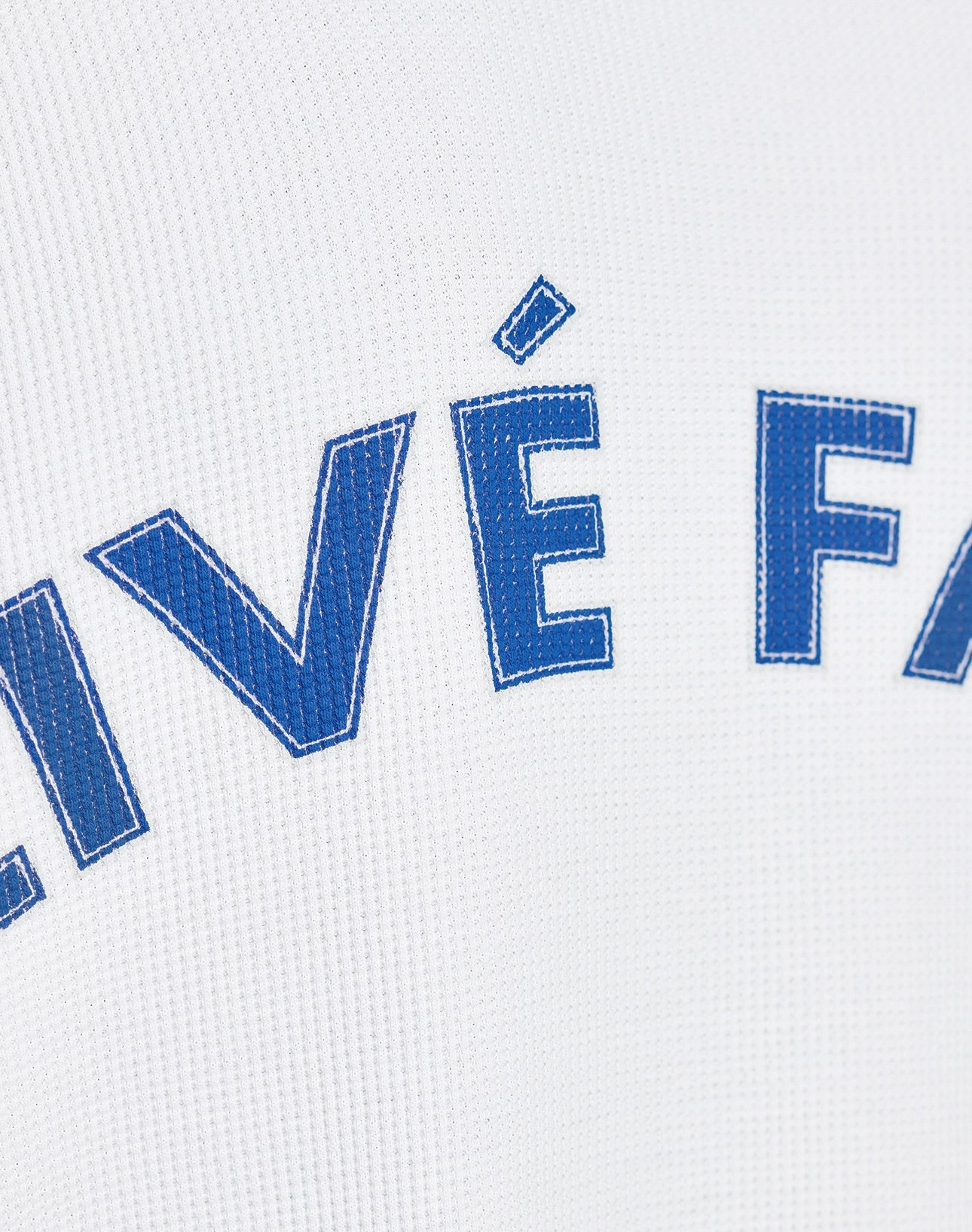 Close-up of blue letters with white stitching on textured white fabric, partially showing VÉ FA. The LÉquipe Waffle Longsleeve by LIVE FAST DIE YOUNG features 100% Baumwolle and an Oversize Fit for extra comfort.