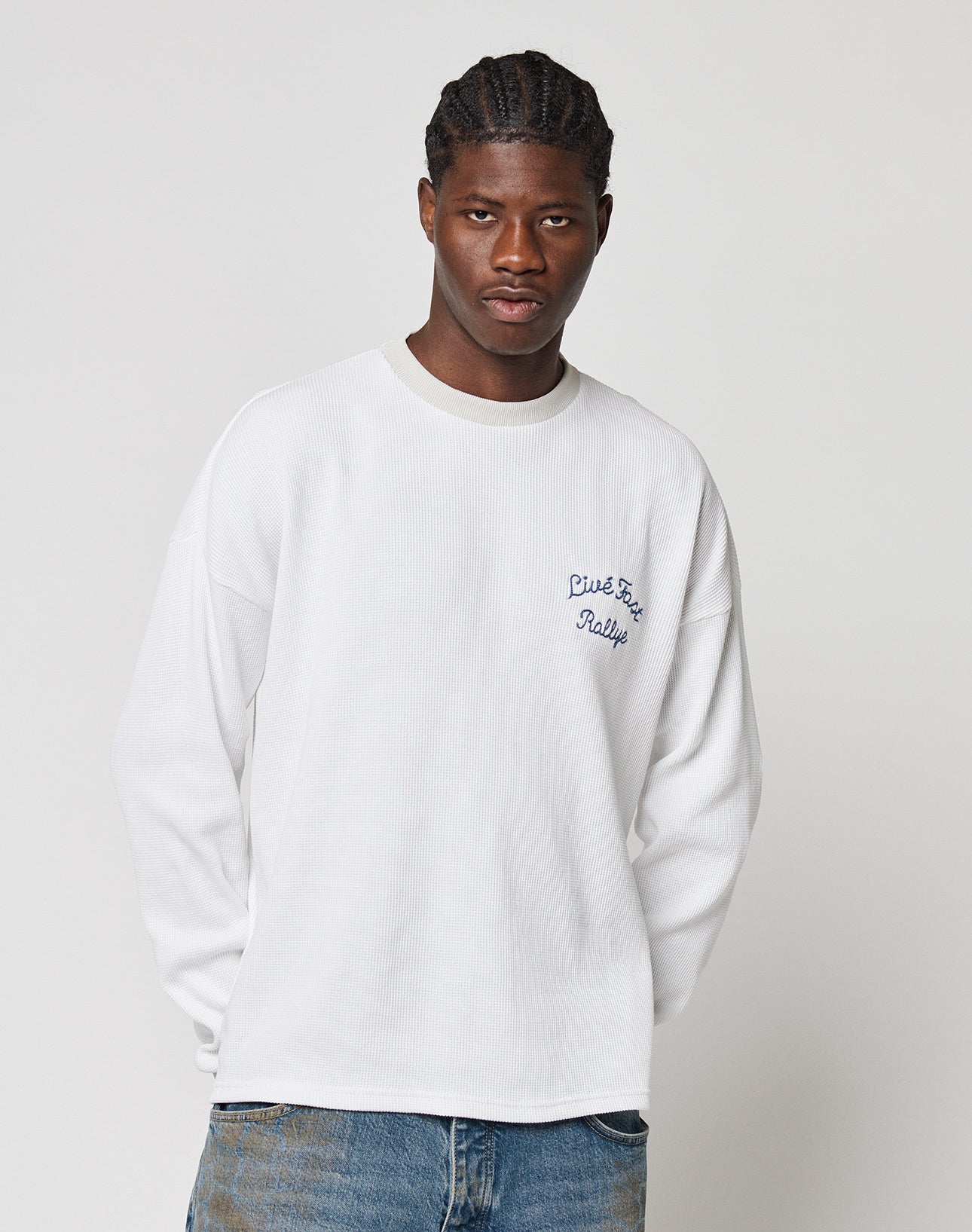 A man with a neutral expression wears the LIVE FAST DIE YOUNG LÉquipe Waffle Longsleeve in white, featuring blue Live Fast Rally embroidery, styled with blue jeans against a plain light gray background.