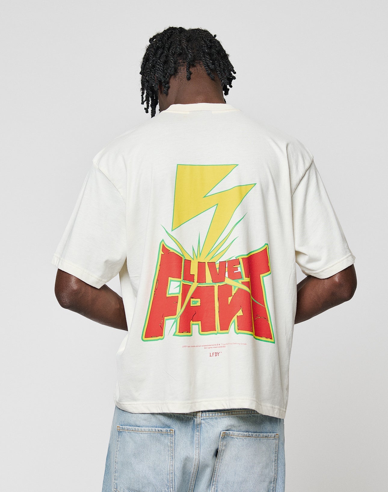 A person with short, twisted hair faces away wearing the LFDY Flash Tee by LIVE FAST DIE YOUNG—an oversized white t-shirt with a bold back graphic: a large yellow lightning bolt over “LIVE FAST” in red and yellow, plus green accents.