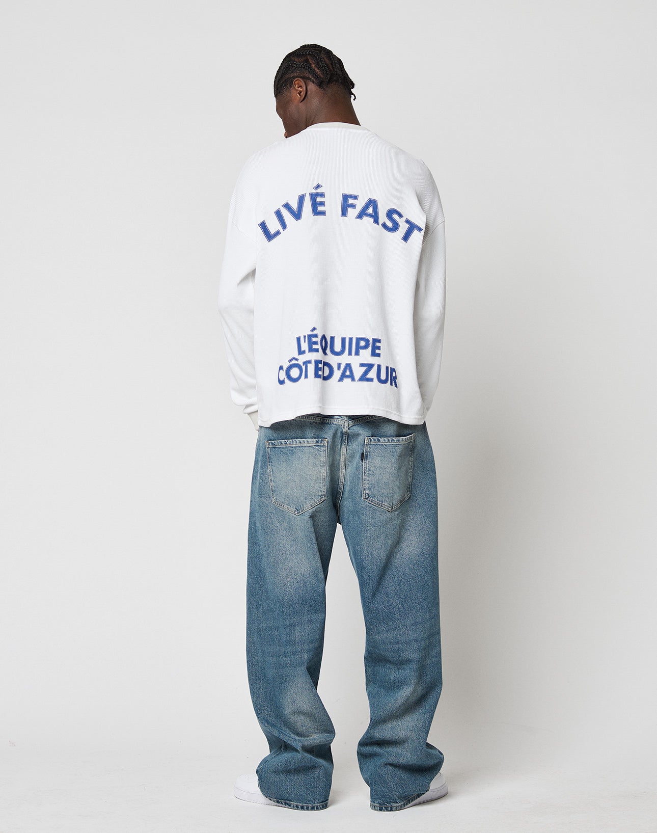 A person faces away in a plain setting, wearing the LIVE FAST DIE YOUNG LÉquipe Waffle Longsleeve—a white oversize sweatshirt with bold blue backprint—paired with baggy blue jeans and white shoes. Crafted from 100% Baumwolle.