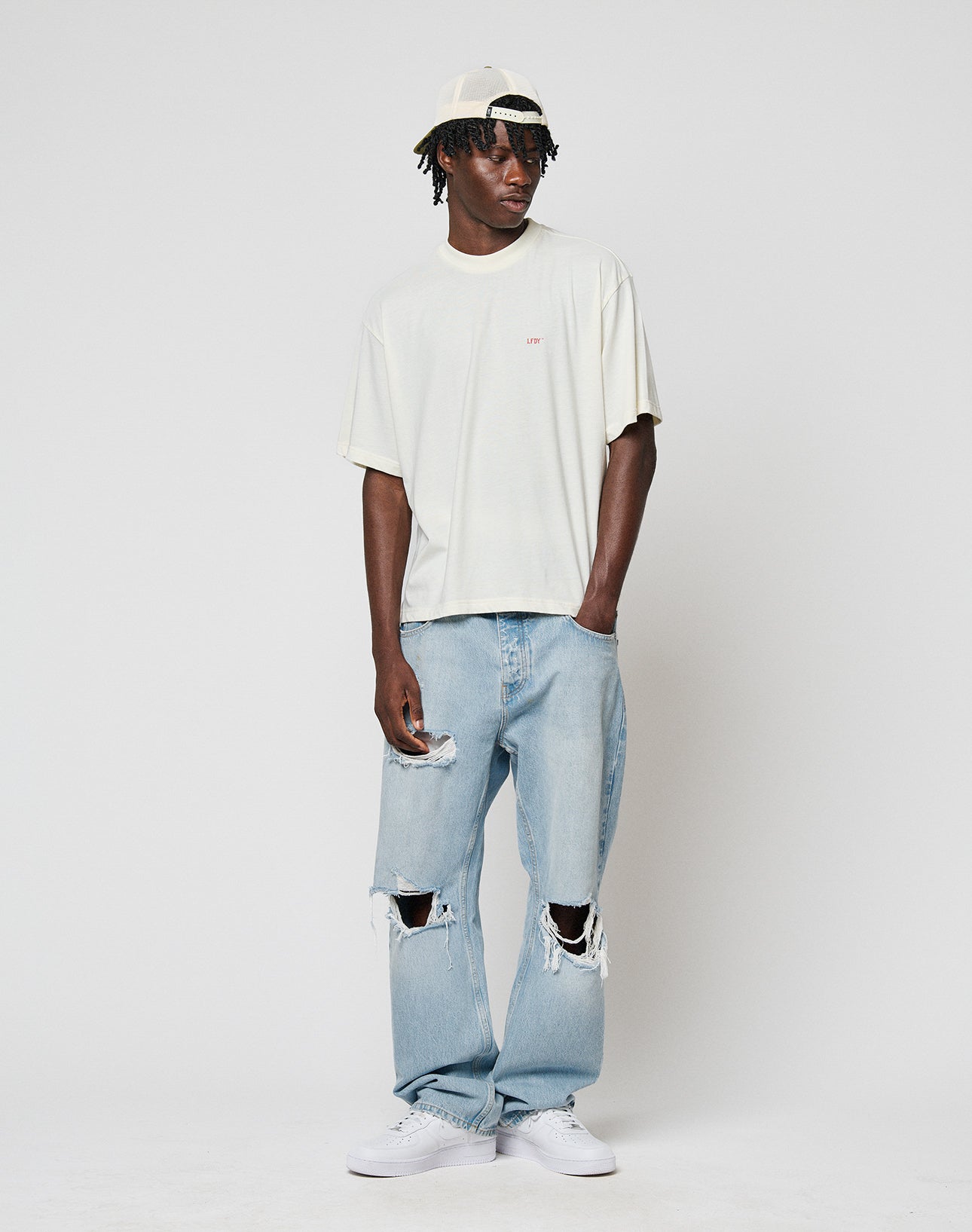 A man wearing a light beige 100% Baumwolle t-shirt, LFDY Distressed Baggy Denim by LIVE FAST DIE YOUNG, white sneakers, and a backwards white cap stands against a white background, looking to the side with one hand in his pocket.