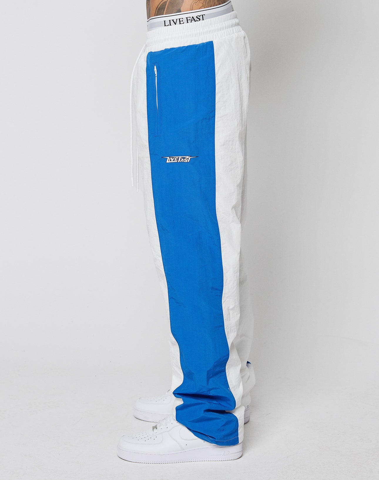 Side view of a person wearing LIVE FAST DIE YOUNG LFDY Trackpants 3000: wide fit, white with bold blue stripe, LIVE FAST on elastic waistband, multi-logo details, zip pocket, LVFST patch—styled with classic white sneakers.