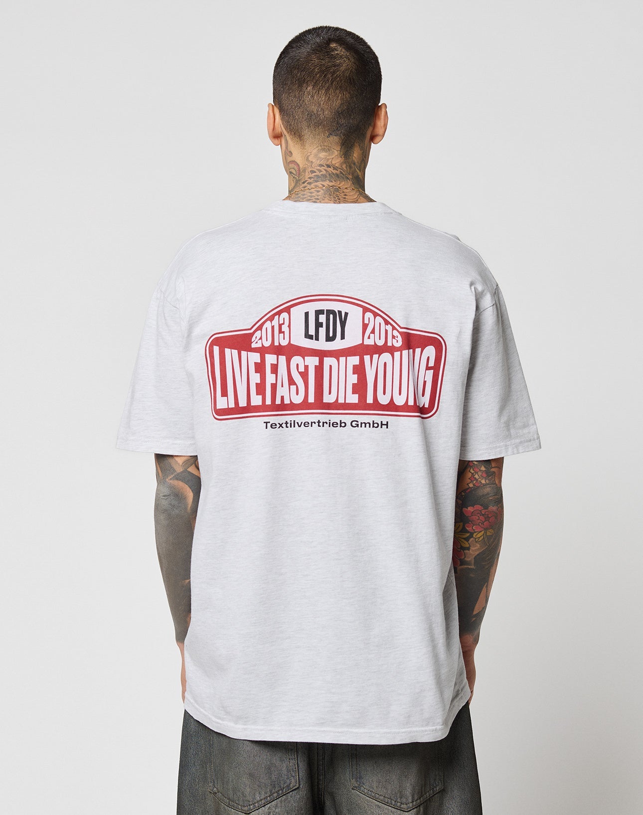 A tattooed person, seen from behind, wears the LIVE FAST DIE YOUNG Live Fast 2013 Tee in light gray with a bold red and black LFDY LIVE FAST DIE YOUNG 2013 graphic on the back.