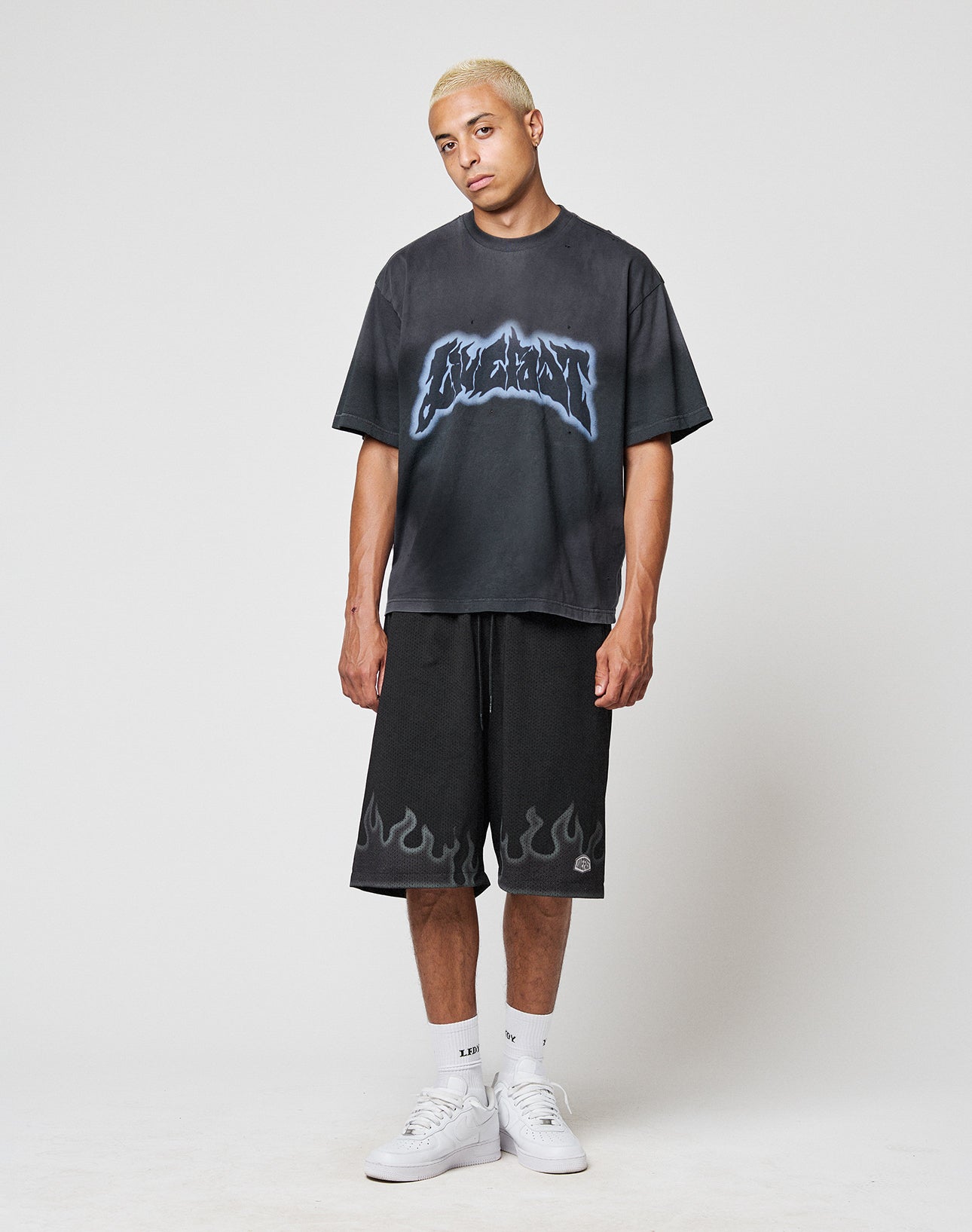 A person with short blond hair wears the LFDY Flames Tee by LIVE FAST DIE YOUNG, featuring blue graphics, paired with black flame shorts, white socks, and white sneakers, standing against a plain white background.