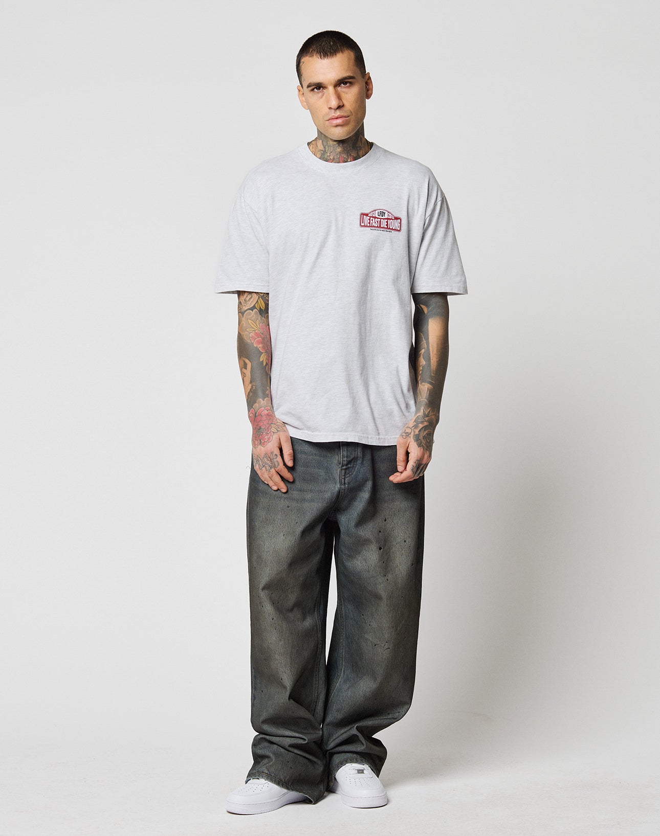 A tattooed man stands against a white background, wearing the LIVE FAST DIE YOUNG Live Fast 2013 Tee aus 100% Baumwolle in Regular Fit, paired with loose dark jeans and white sneakers. His arms are relaxed by his sides and he has a neutral expression.