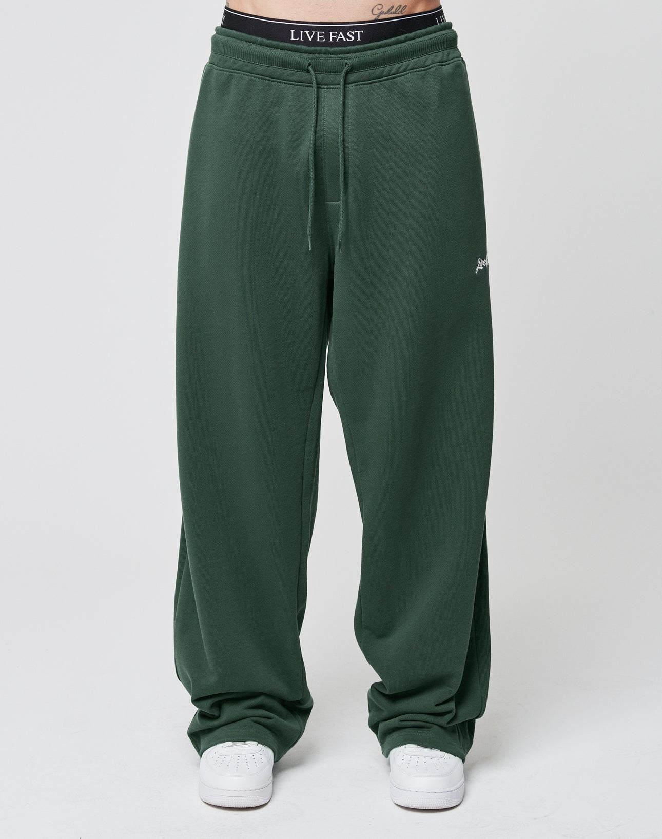 A person wears loose-fitting, dark green Basic Wide Jogger pants by LIVE FAST DIE YOUNG with a wide fit and elastic waistband, white sneakers, and black LIVE FAST underwear visible above the waistband. The background is plain and light-colored.