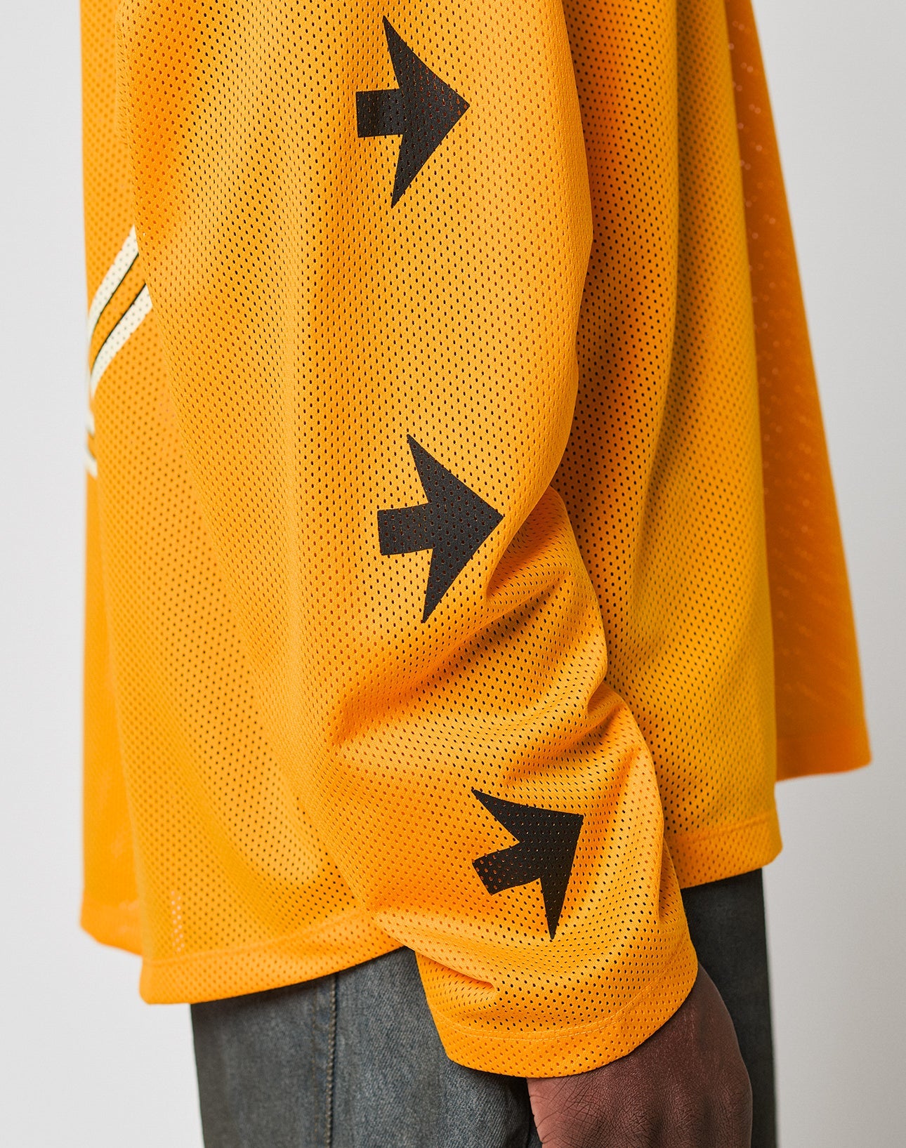 A person wears the LFDY Autobahn Jersey by LIVE FAST DIE YOUNG—an orange oversized polyester mesh long-sleeve shirt with three black arrow symbols on the sleeve—paired with dark pants against a plain light background.