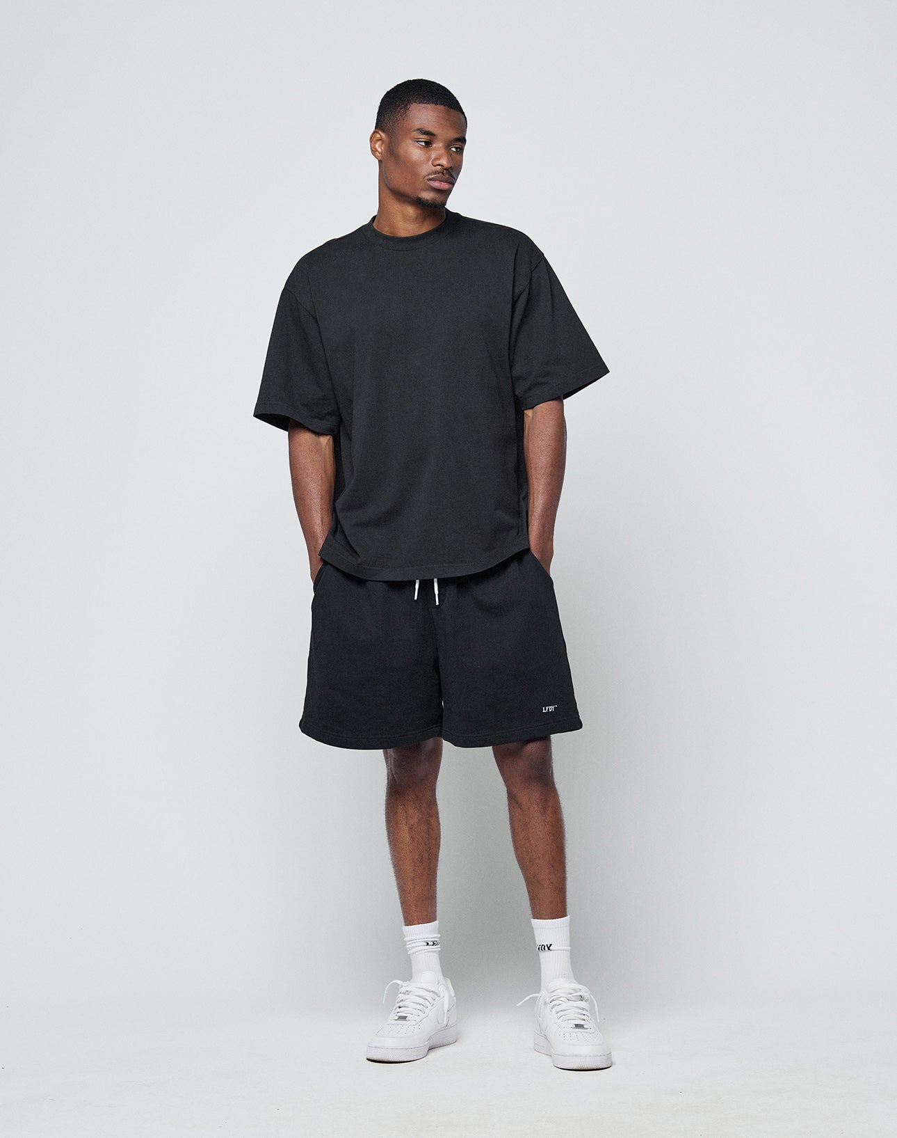 A man stands against a plain light gray background wearing the LIVE FAST DIE YOUNG Basic Heavy Tee in black, an oversize 100% Baumwolle T-shirt, paired with black drawstring shorts, white crew socks, and white sneakers. His hands are in his pockets.