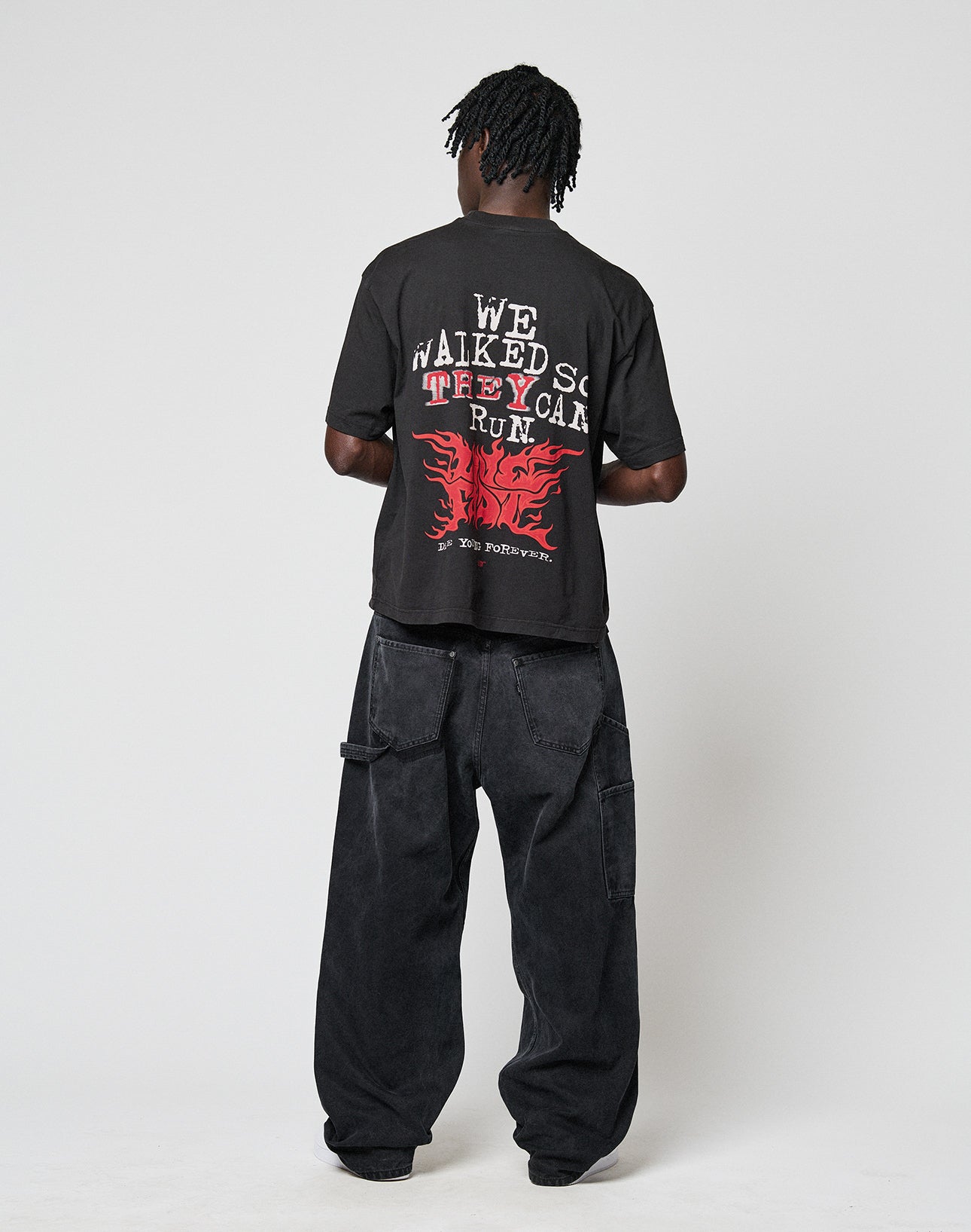 A person faces away, wearing the LFDY We Walked Tee by LIVE FAST DIE YOUNG—an oversized black T-shirt with bold white and red front and back graphics—paired with loose black jeans against a plain light background.