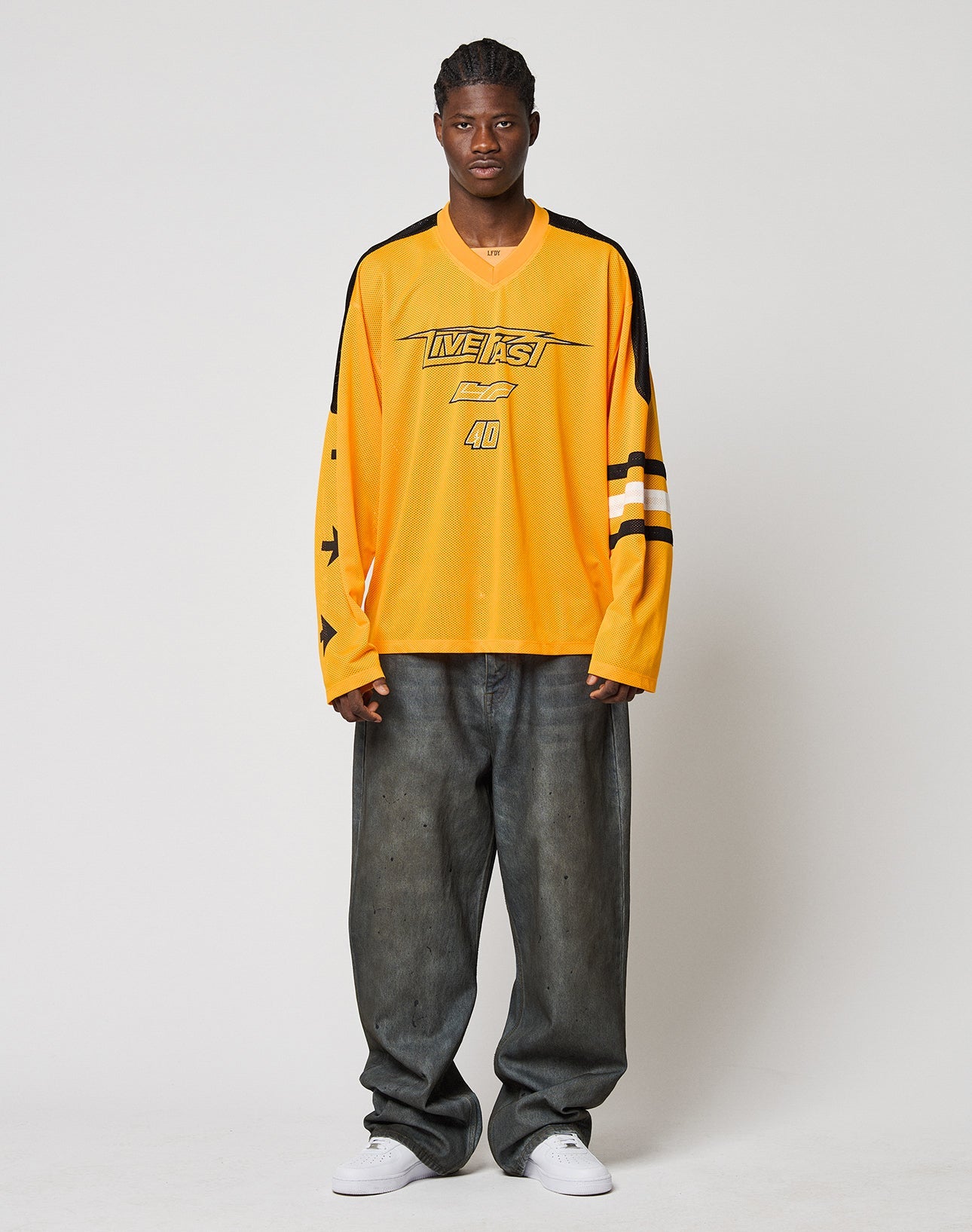 A young man stands against a plain background, wearing the LIVE FAST DIE YOUNG LFDY Autobahn Jersey in yellow with black and white details, paired with oversized faded gray jeans and white sneakers.