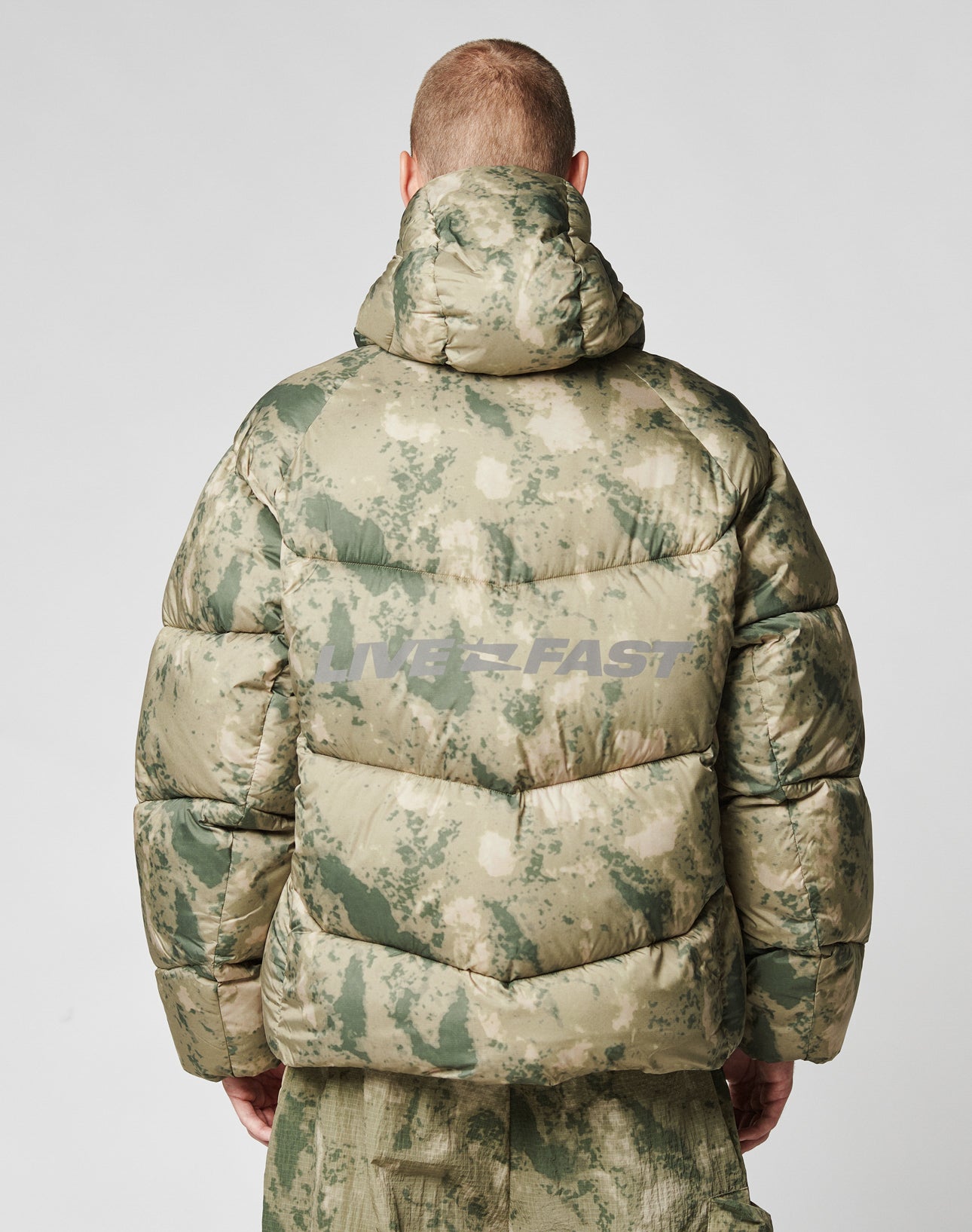 A person seen from behind wears the LIVE FAST DIE YOUNG Live Fast Puffer Jacket in green and beige camo with a hood and a subtle reflective WESTFAST backprint, paired with matching camouflage pants.
