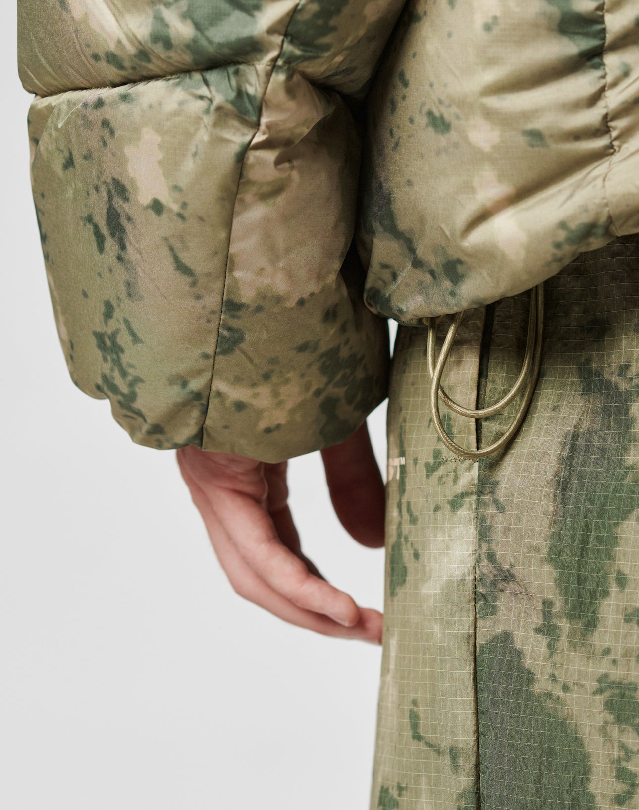 Close-up of a persons hand by their side, wearing the LIVE FAST DIE YOUNG Live Fast Puffer Jacket in green and beige camouflage with an oversized fit and drawstrings at the waist, paired with matching pants.