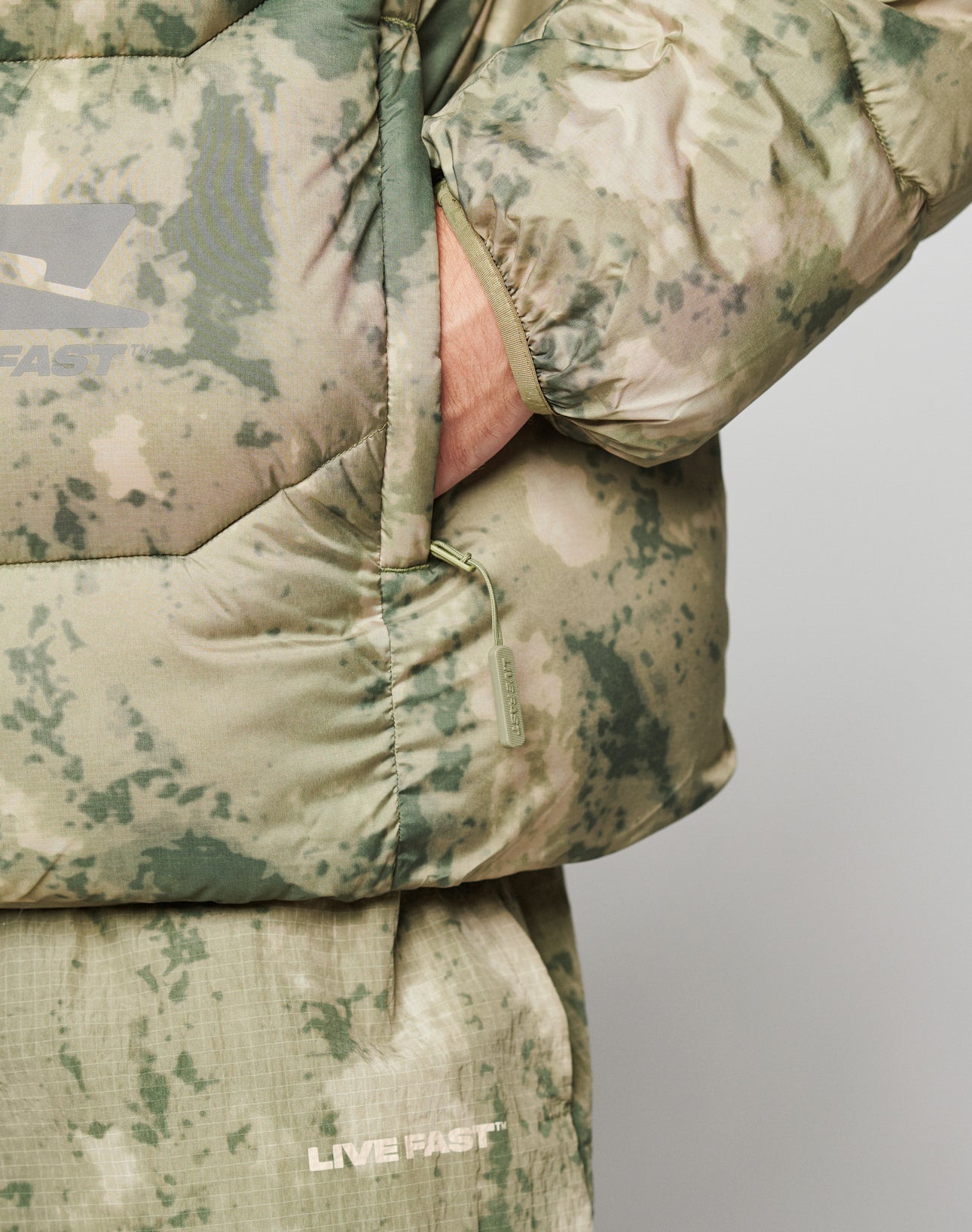 A close-up of a hand in the pocket of the green camouflage Live Fast Puffer Jacket by LIVE FAST DIE YOUNG, featuring “LIVE FAST” as a reflective front and back print in white.
