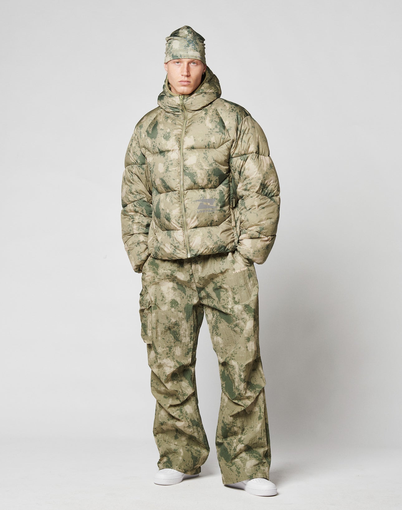 A person faces forward in an oversize green and beige camo puffer with elastikkordeln, matching Live Fast Nylon Pants by LIVE FAST DIE YOUNG with a practical cargotasche, a camo cap, and white sneakers against a plain light gray background.