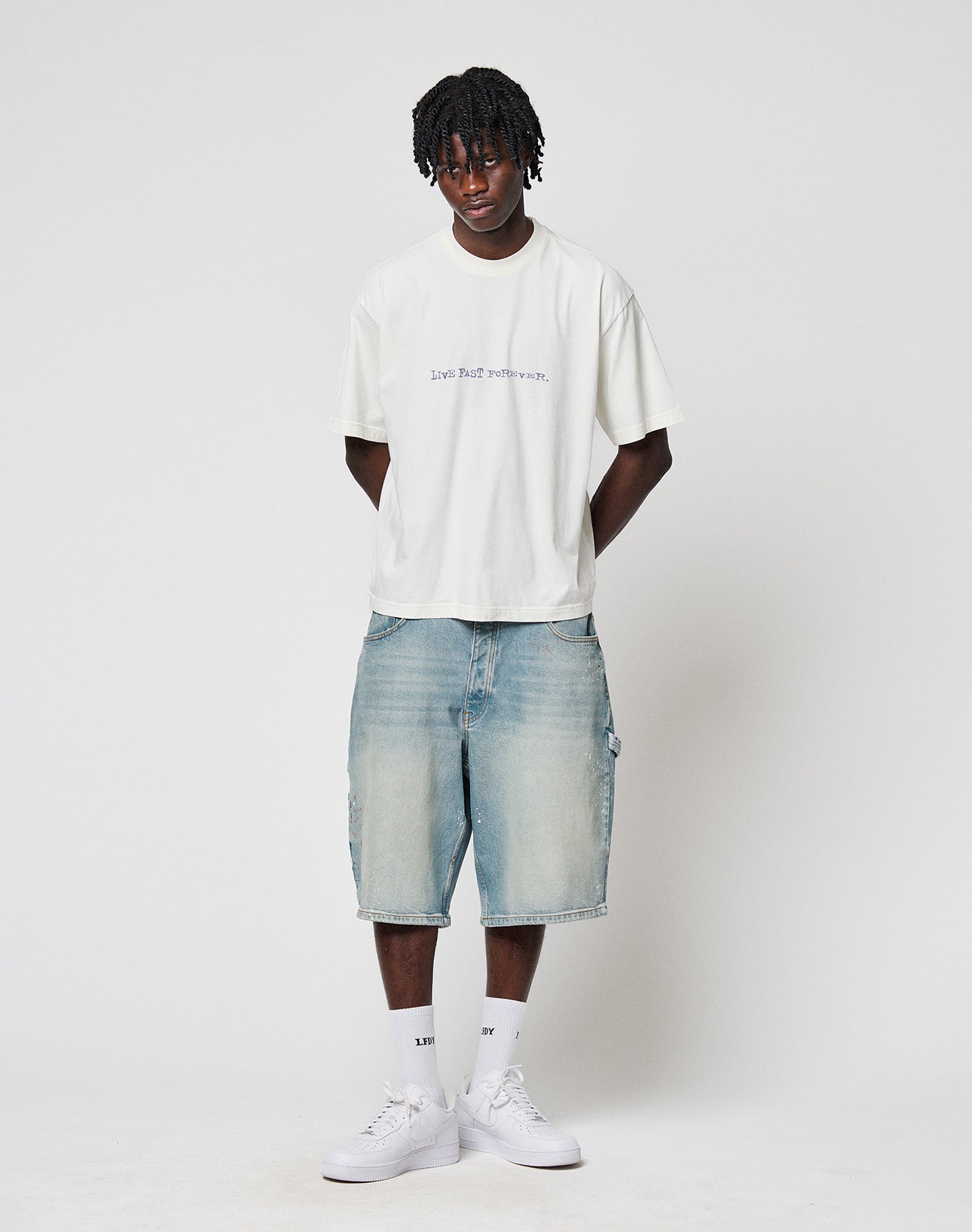 A young man with short dreadlocks poses against a white background, wearing the oversize LFDY We Walked Tee by LIVE FAST DIE YOUNG with front and back print, light blue knee-length denim shorts, white socks, and white sneakers.