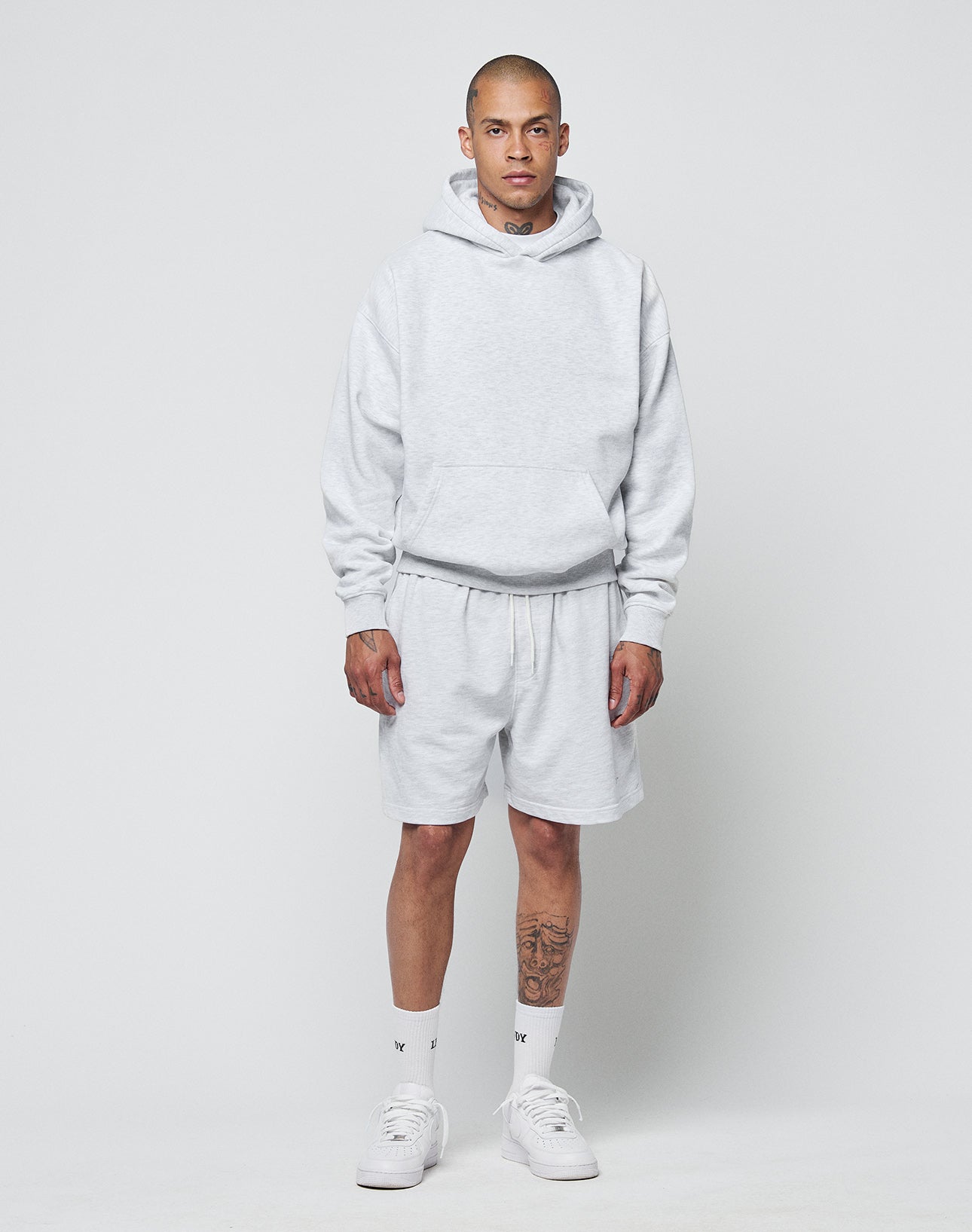 A person faces forward wearing an oversize fit, light gray LIVE FAST DIE YOUNG Basic Blank Hooded hoodie and matching shorts, with white crew socks, white sneakers, and a tattoo on their left calf. The background is plain and light.