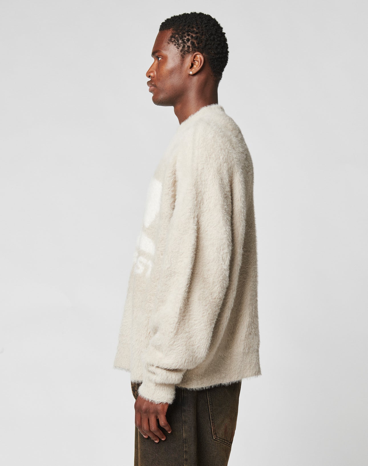 A man in profile faces left against a plain background, wearing the LIVE FAST DIE YOUNG Live Fast Fuzzy Sweater in oversize light beige with dark pants. His arms are relaxed at his sides.