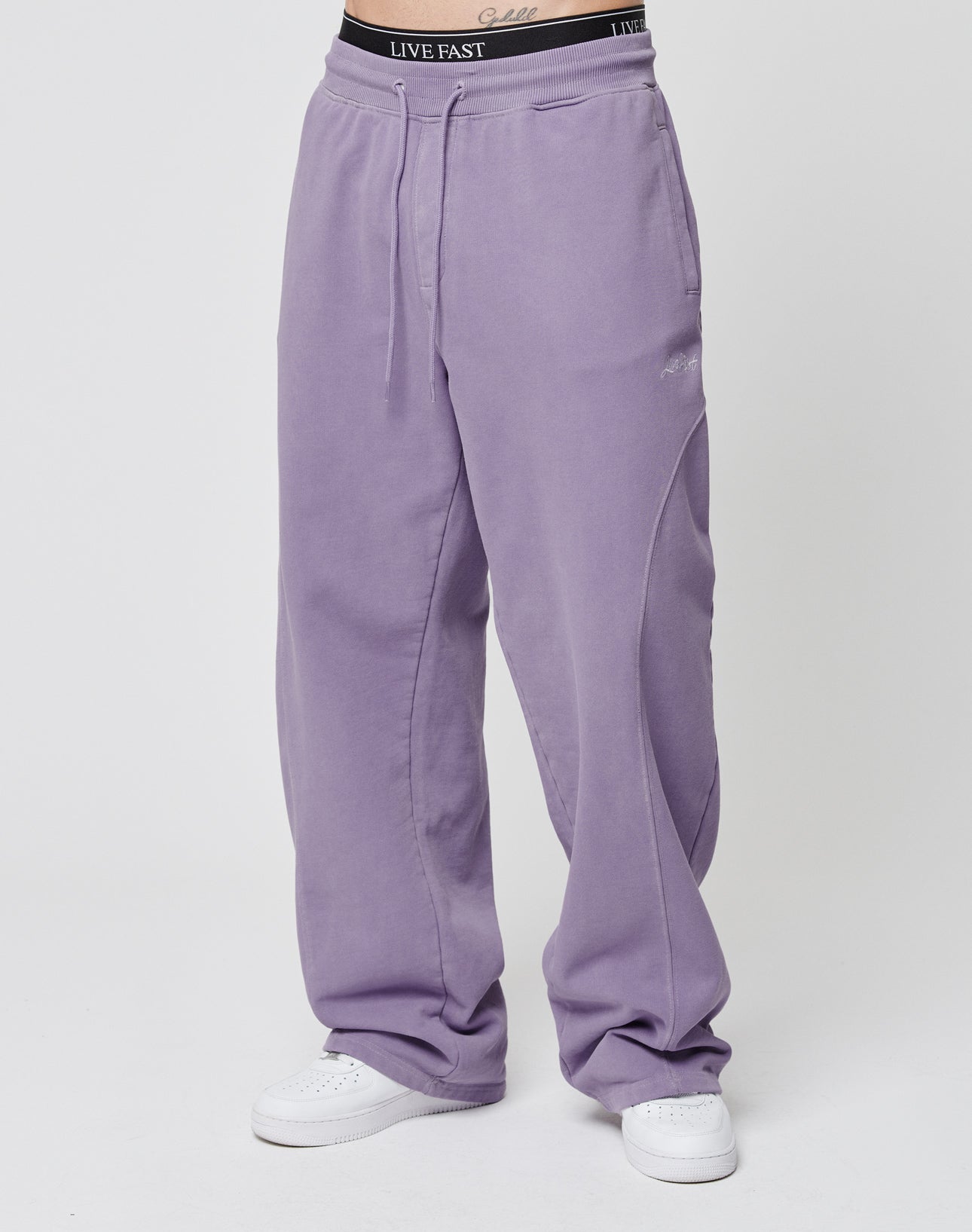 A person wears loose-fitting lavender Basic Wide Jogger sweatpants by LIVE FAST DIE YOUNG with an elastic waistband and drawstrings, white sneakers, and a visible black waistband that reads LIVE FAST on a plain white background.