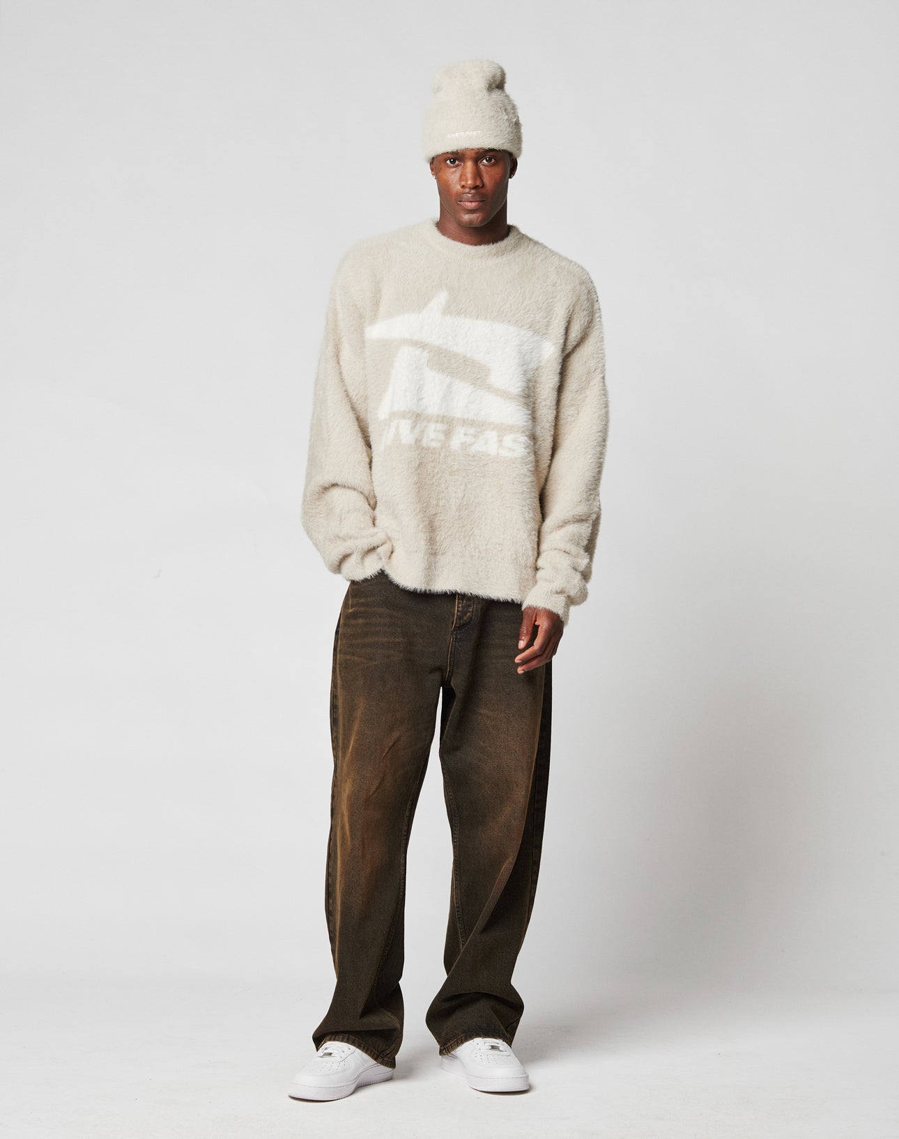 A man stands against a plain white background wearing the LIVE FAST DIE YOUNG Live Fast Fuzzy Sweater in beige with a white intarsia logo and text, paired with loose dark brown jeans, white sneakers, and a matching beige beanie.