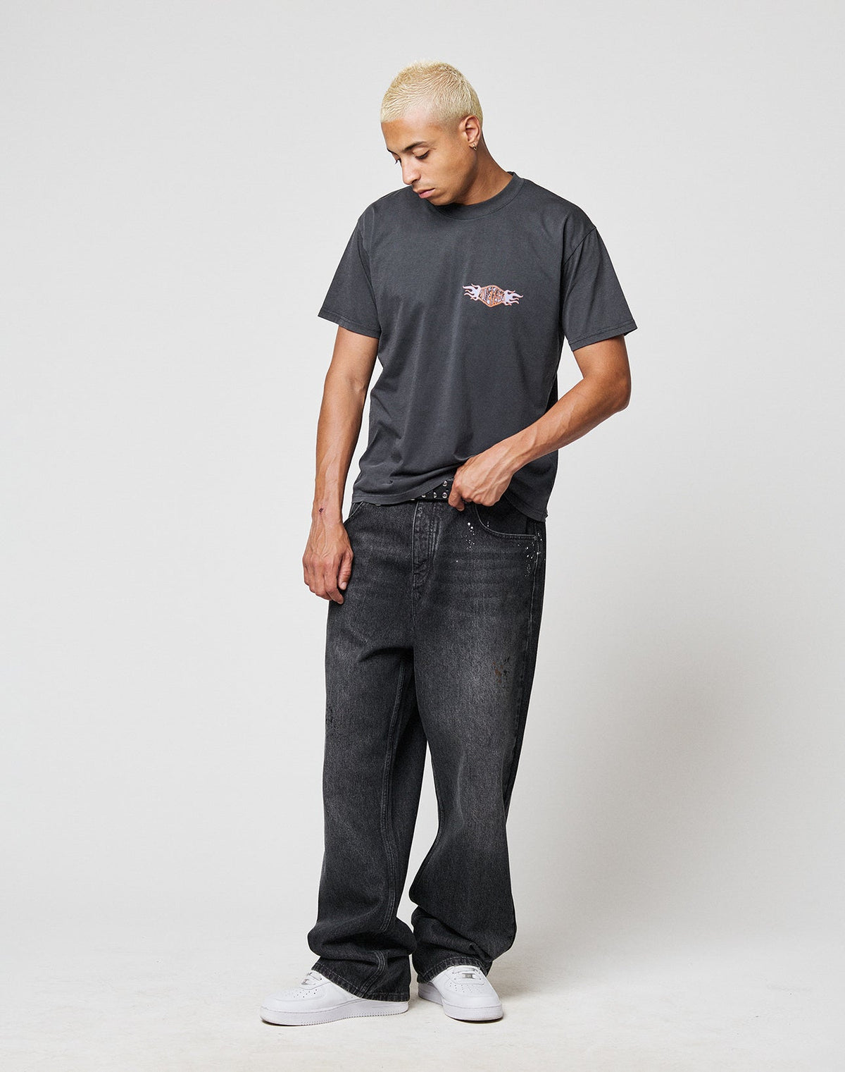 A person with short blond hair wears a loose dark gray t-shirt with a small chest graphic, LFDY Baggy Denim by LIVE FAST DIE YOUNG, and white sneakers, standing against a plain white background and looking down.