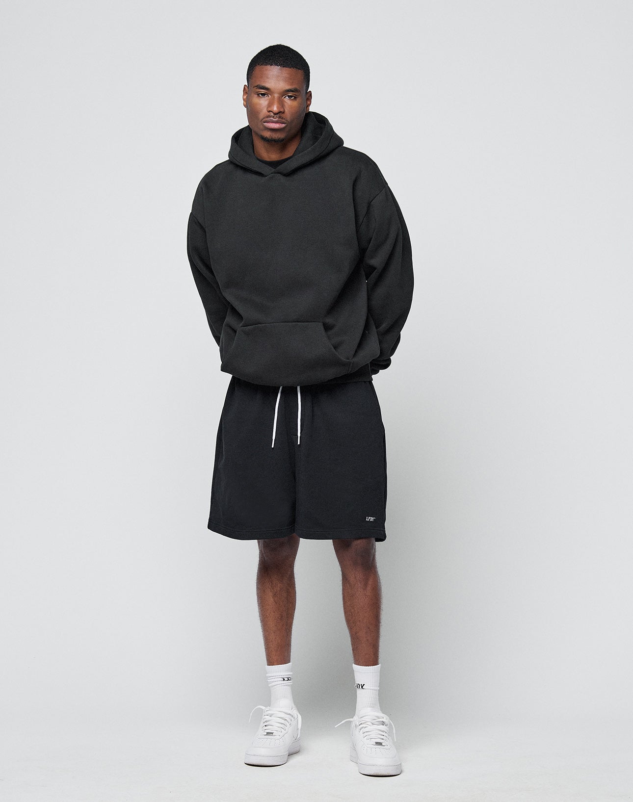 A man stands against a light grey background wearing the LIVE FAST DIE YOUNG Basic Blank Hooded black hoodie, black shorts with a white drawstring, white socks, and white sneakers. His hands are behind his back as he looks at the camera.