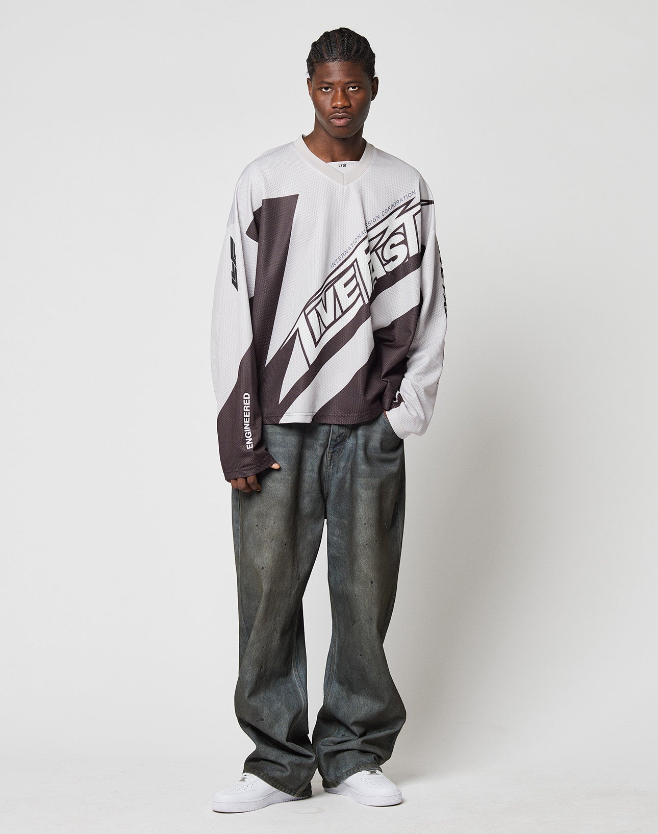 A man with braided hair and white sneakers stands against a plain white background, wearing a long-sleeve graphic shirt and LFDY Coated Baggy Denim jeans from LIVE FAST DIE YOUNG.