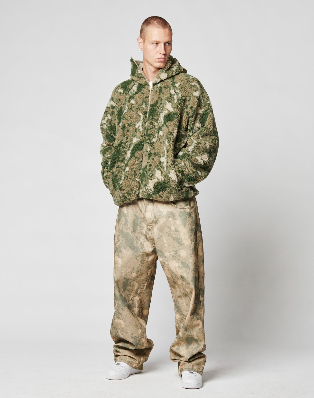 A person stands against a plain background wearing the LIVE FAST DIE YOUNG Live Fast Teddy Jacket in green camouflage with Kapuze mit Visor-Öffnung, loose beige camo pants, and white sneakers, hands in pockets and looking to the side.