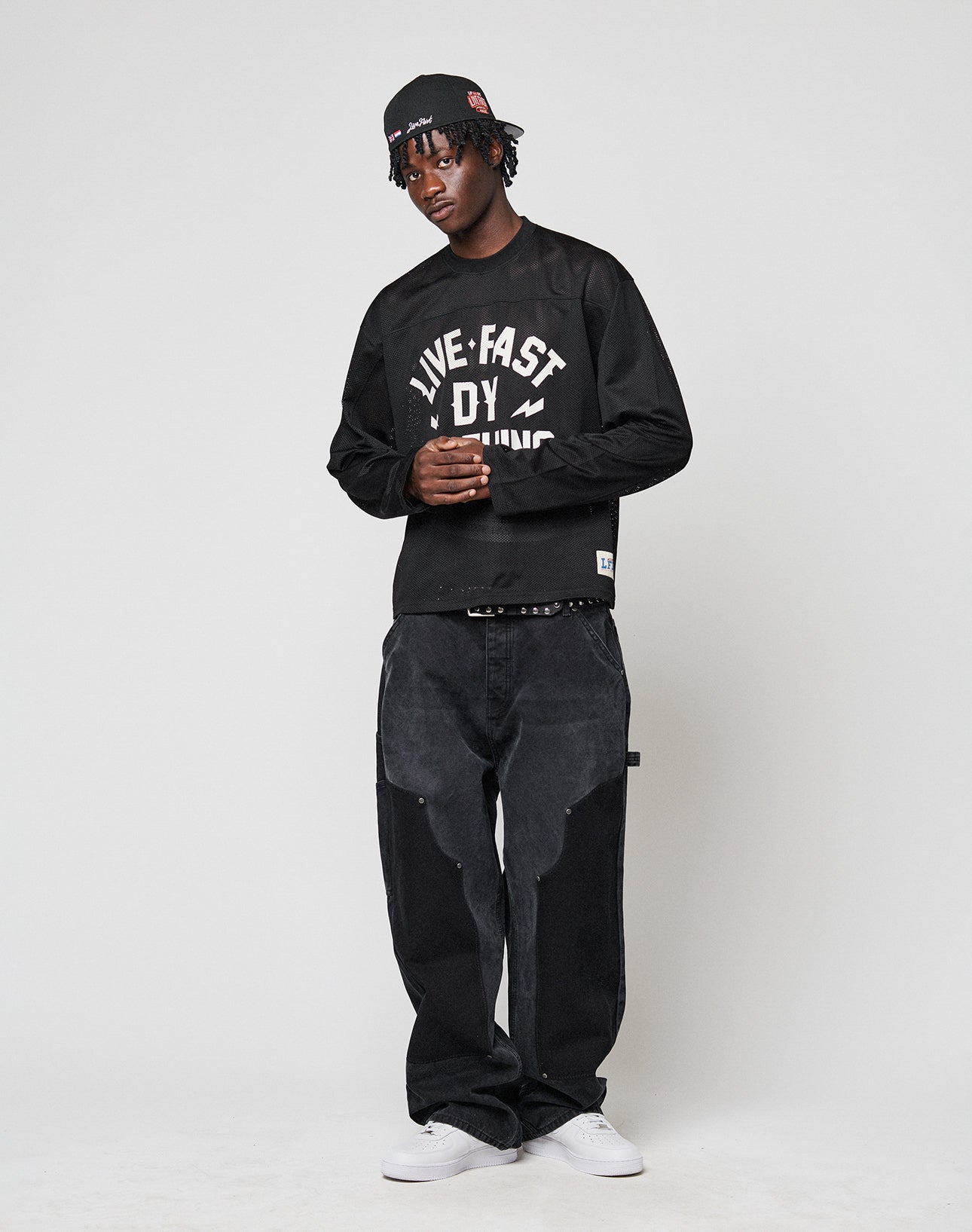 A young man wears the LFDY Flash Jersey by LIVE FAST DIE YOUNG—an oversize black long-sleeve with LIVE FAST frontprint—paired with loose black jeans, black cap, and white sneakers as he stands relaxed against a plain light background.