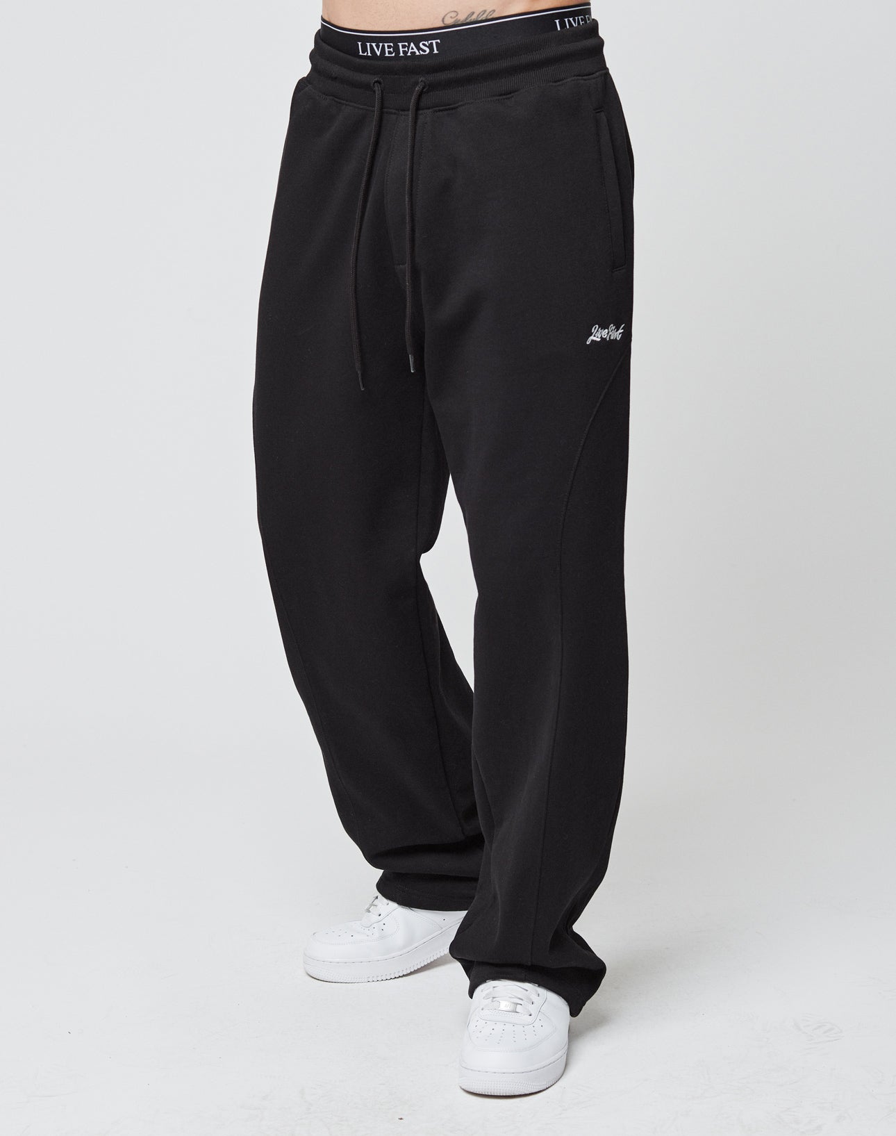 Someone stands against a plain white background wearing LIVE FAST DIE YOUNG’s Basic Wide Jogger in black (wide fit, elastic waistband), white sneakers, and black underwear with ‘LIVE FAST’ on the waistband.