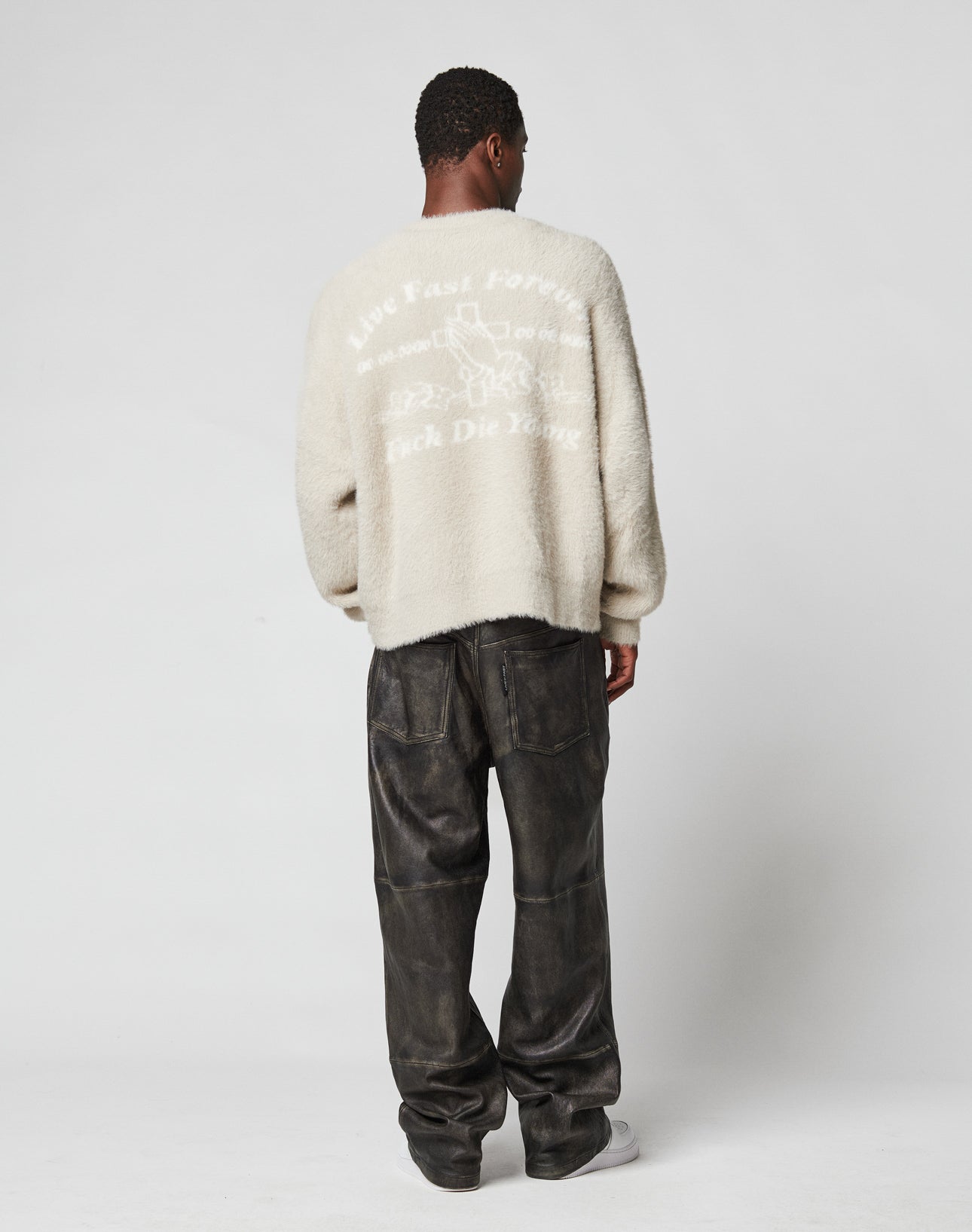 A person faces away in a cream LFDY Praying Fuzzy Sweater by LIVE FAST DIE YOUNG, featuring an oversize fit and intarsia logo on the back, rundhals neckline, and paired with loose dark leather pants against a plain white background.